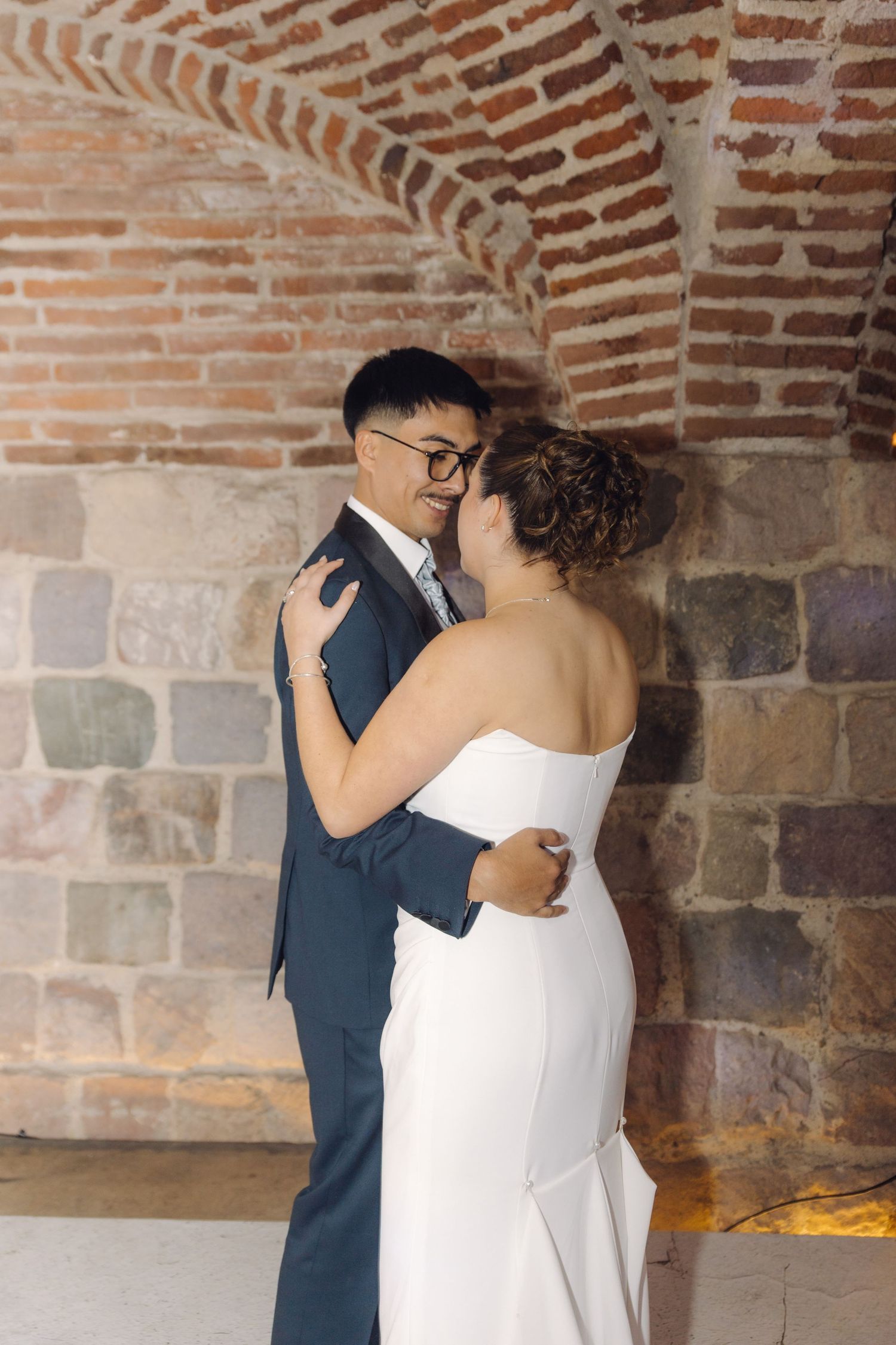 Newlyweds share an intimate embrace while dancing under rustic brick arched walls at their wedding.