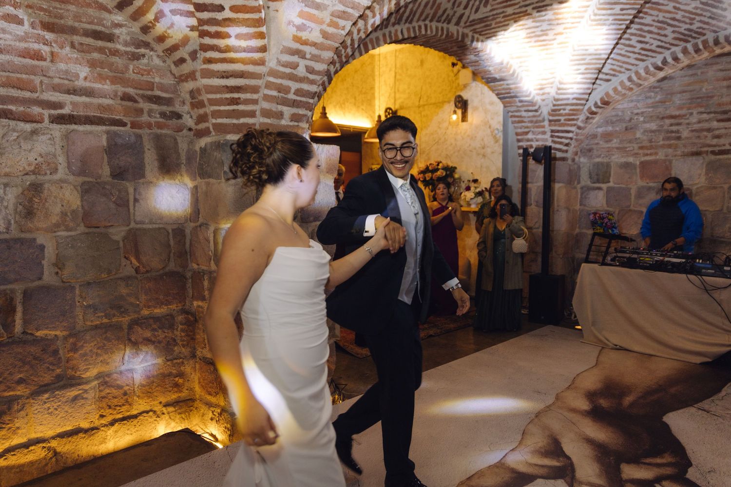 Smiling bride and groom dance joyfully under brick vaulted ceiling at romantic wedding reception.