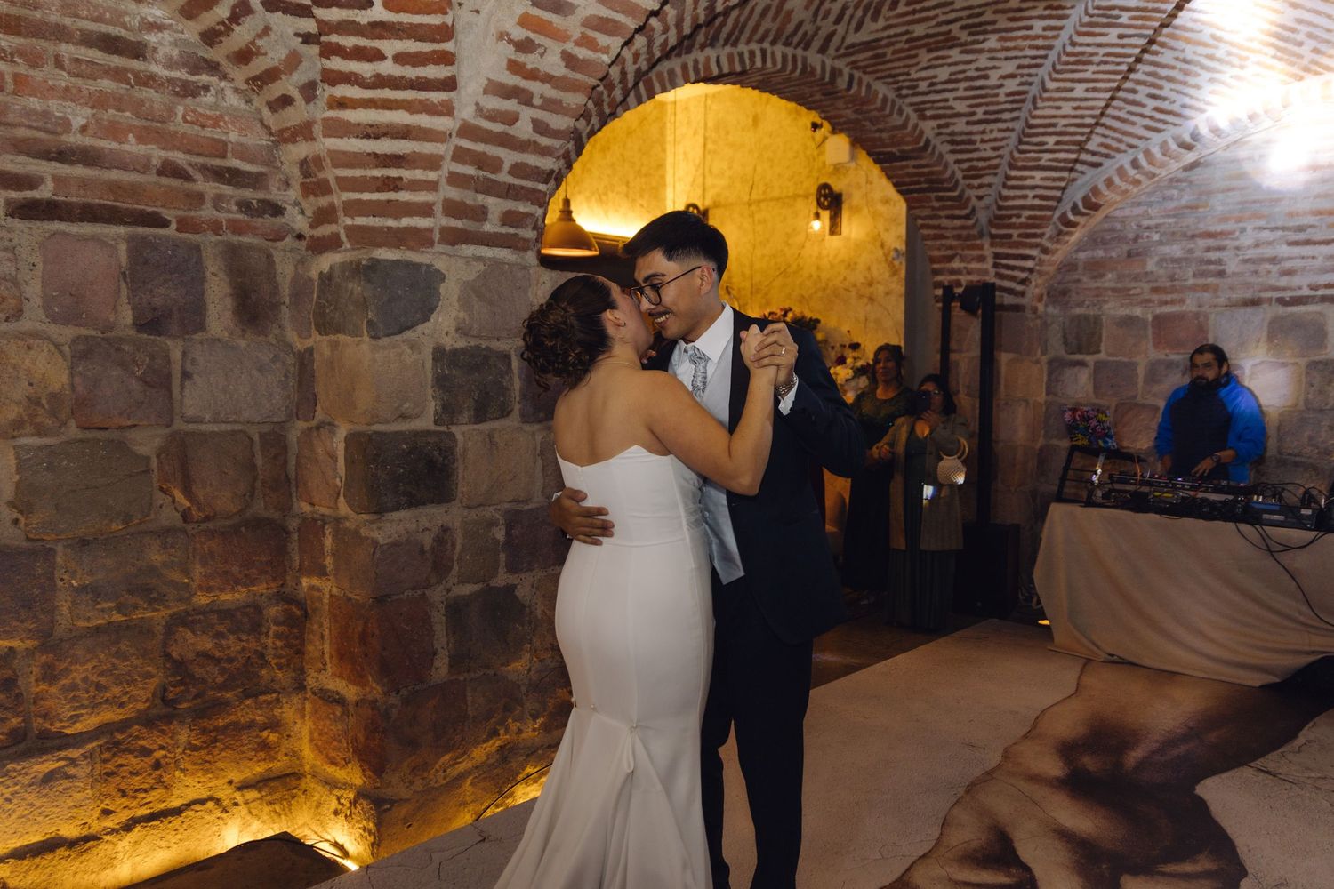 Couple shares romantic first dance embrace in warmly lit brick vaulted wedding reception venue.