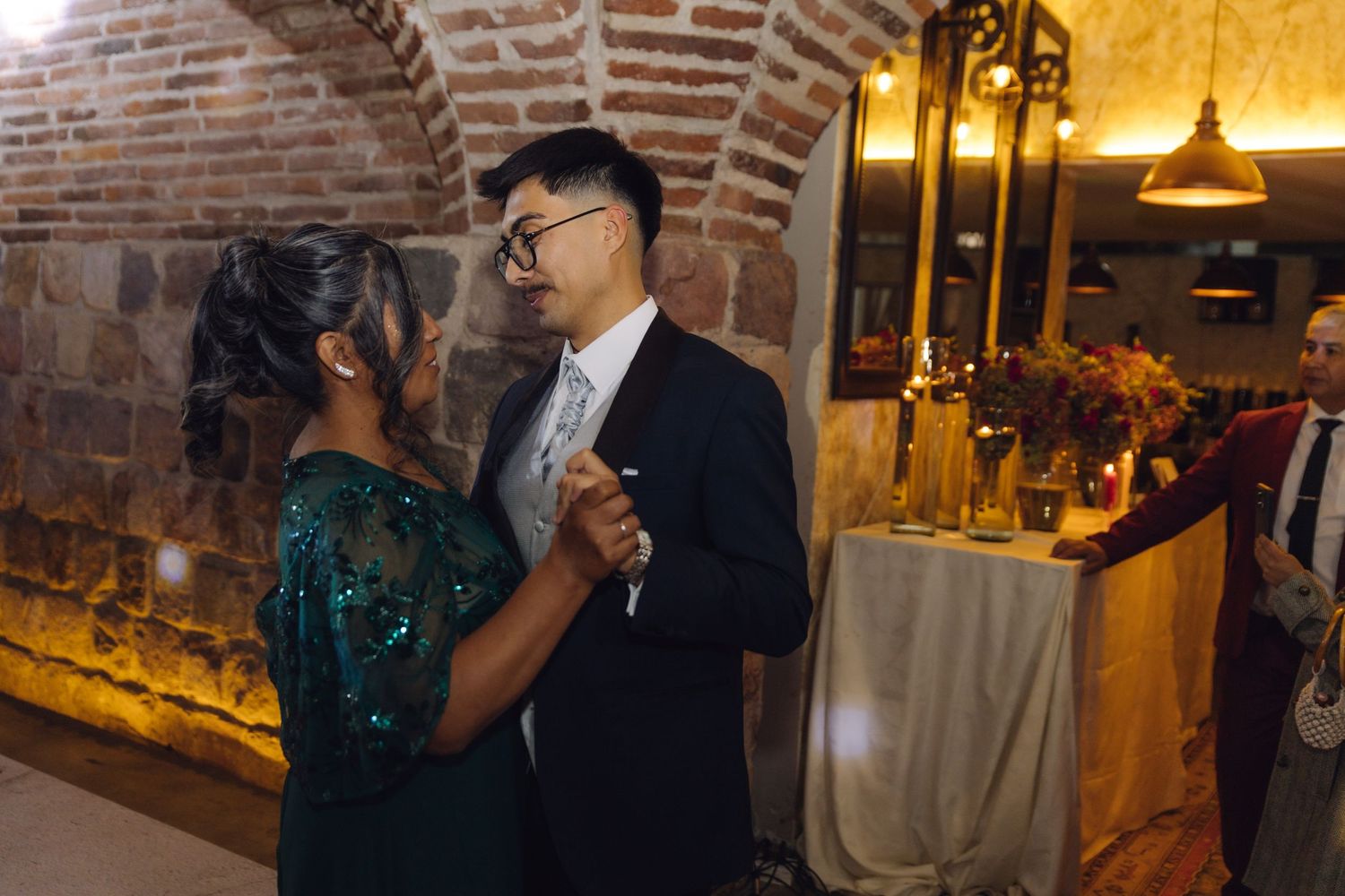 Groom in navy suit dances with woman in green sequin dress at wedding celebration.