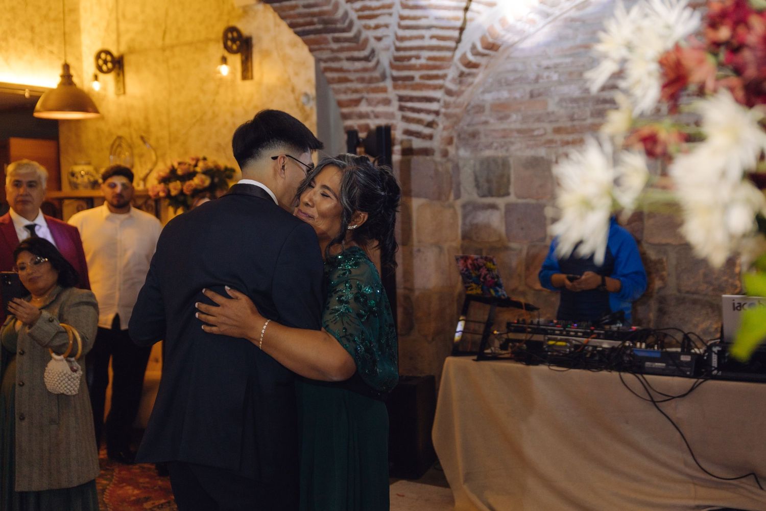 Groom embraces woman in green dress on dance floor at warmly lit wedding reception.