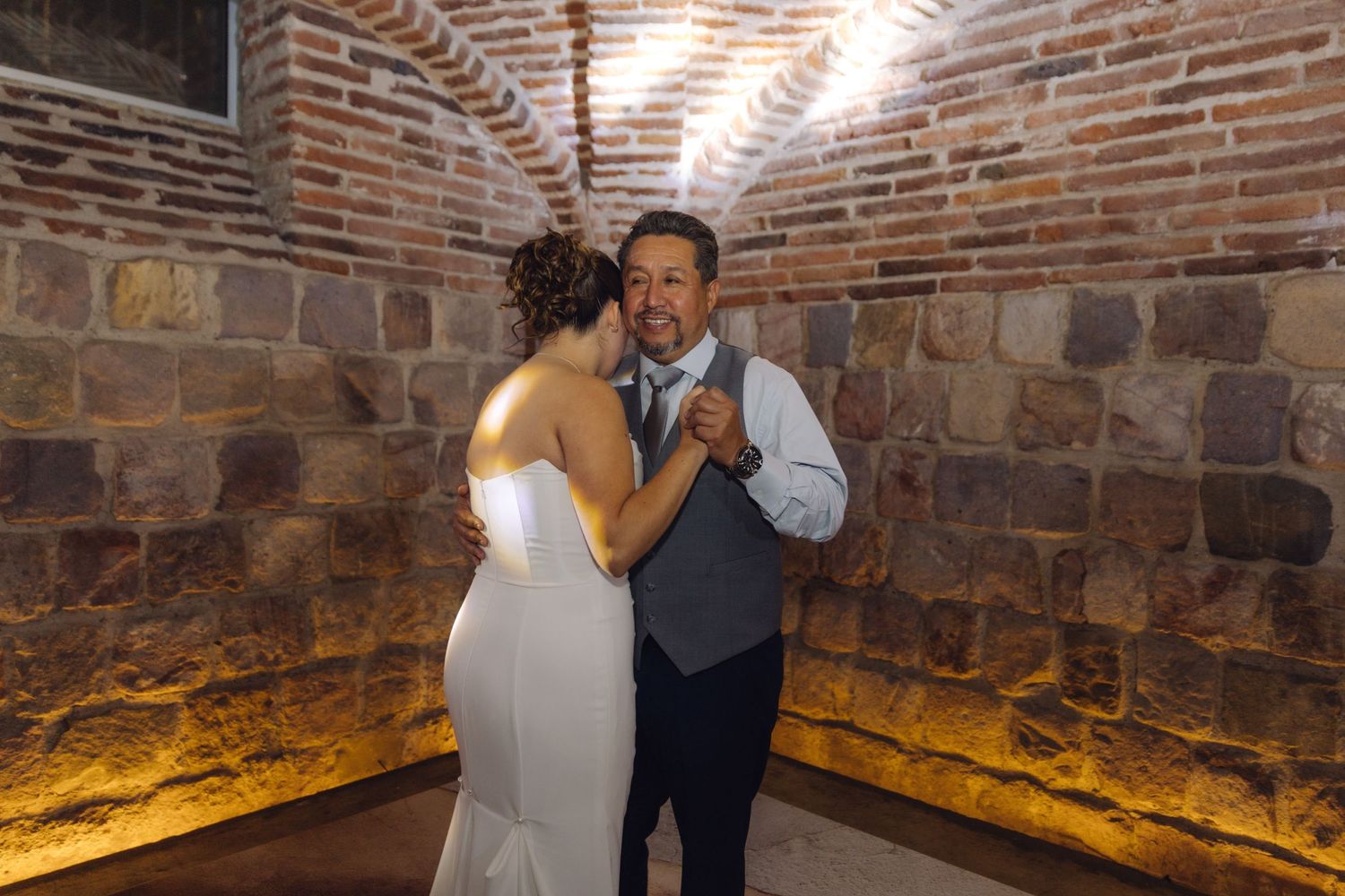 Bride in white gown dances with smiling man in vest under brick arched venue.