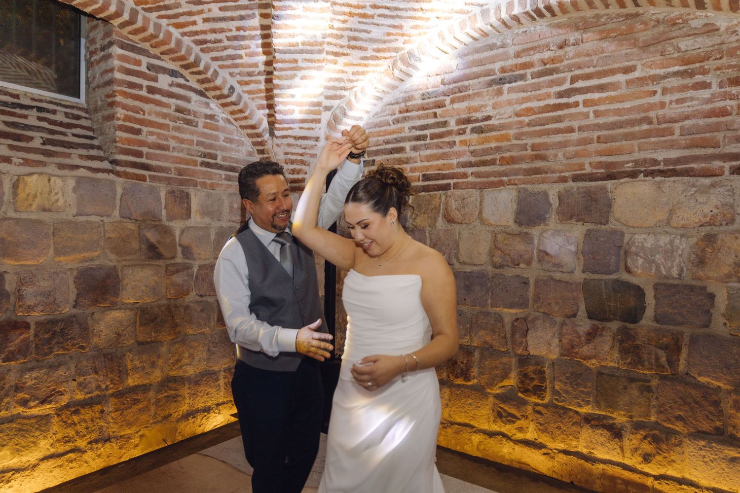 Groom raises bride's hand in a graceful spin during first dance at rustic venue.