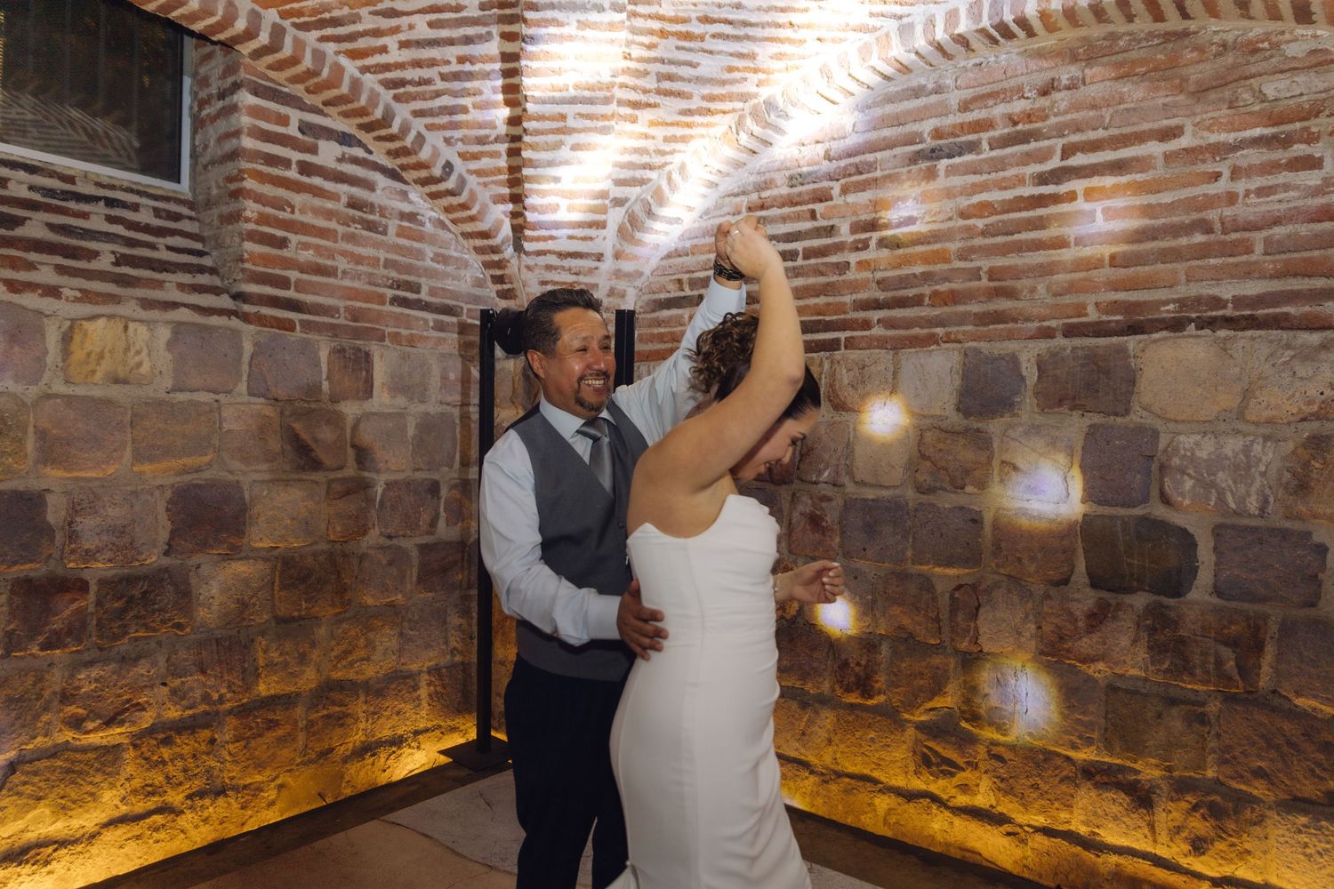 Groom twirls bride in white wedding gown under sparkling lights in brick-walled reception hall.