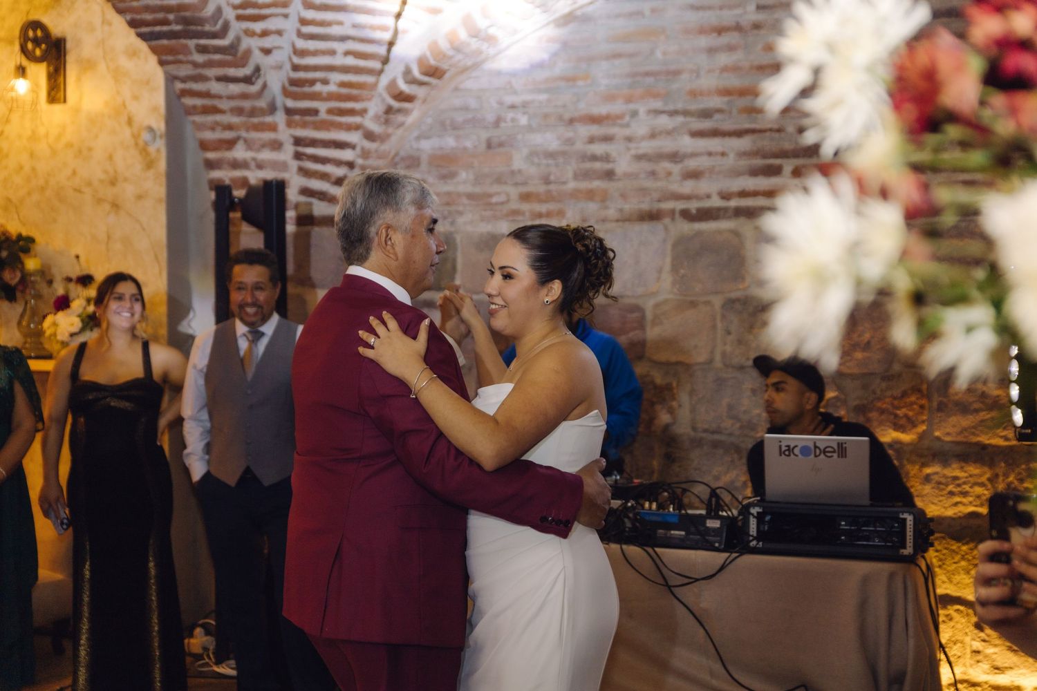 Bride in white gown shares a dance with older man in burgundy suit at reception.