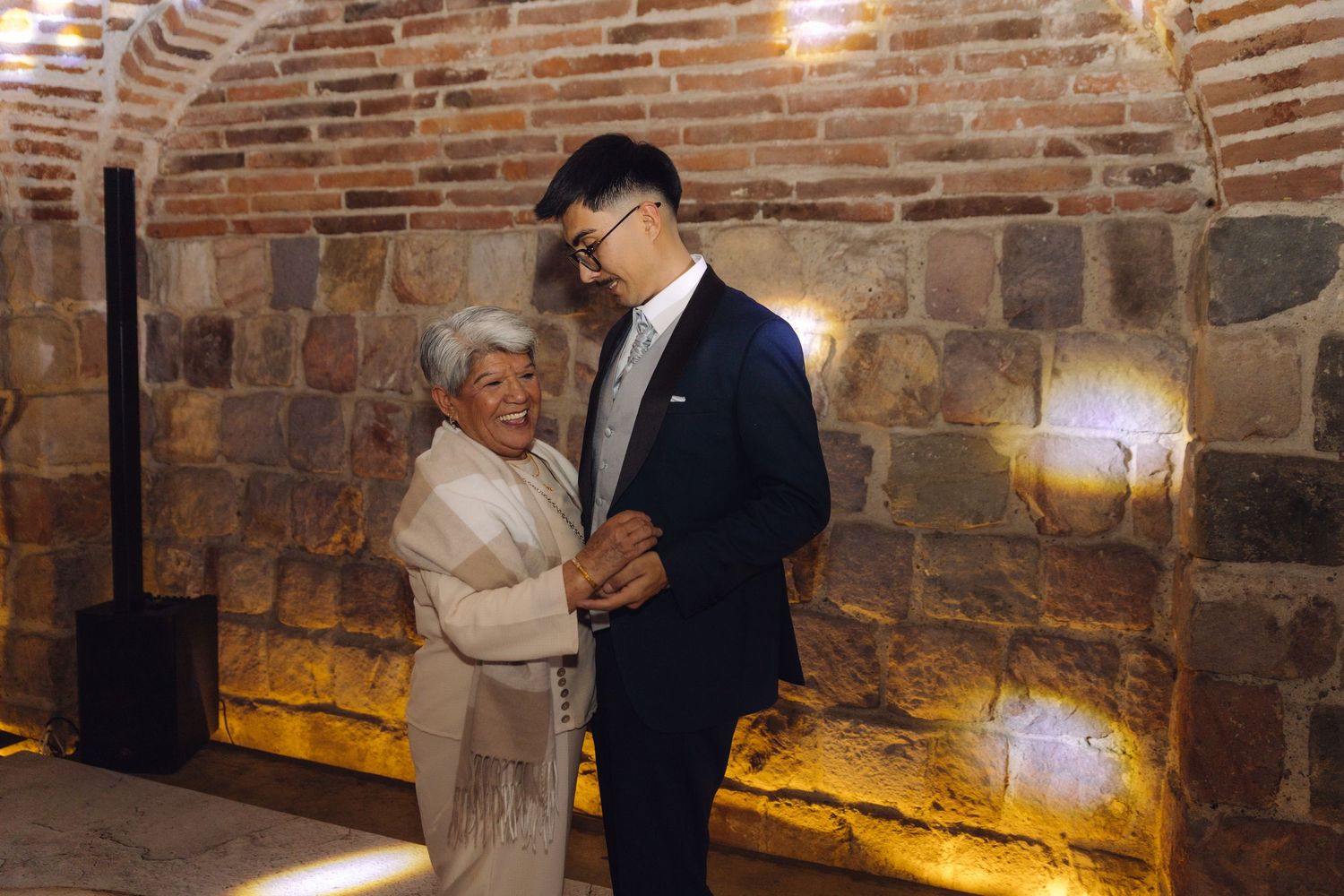 Groom dancing with elderly woman in elegant attire at a wedding celebration indoors.