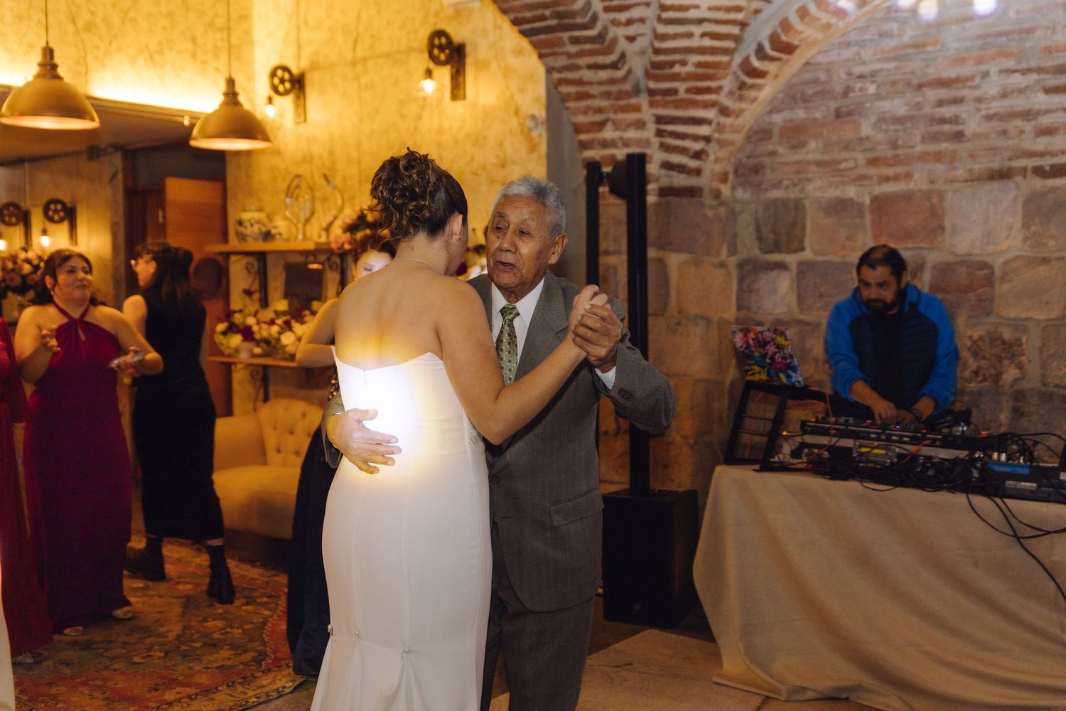 Bride in white gown shares father-daughter dance at rustic wedding venue with DJ.