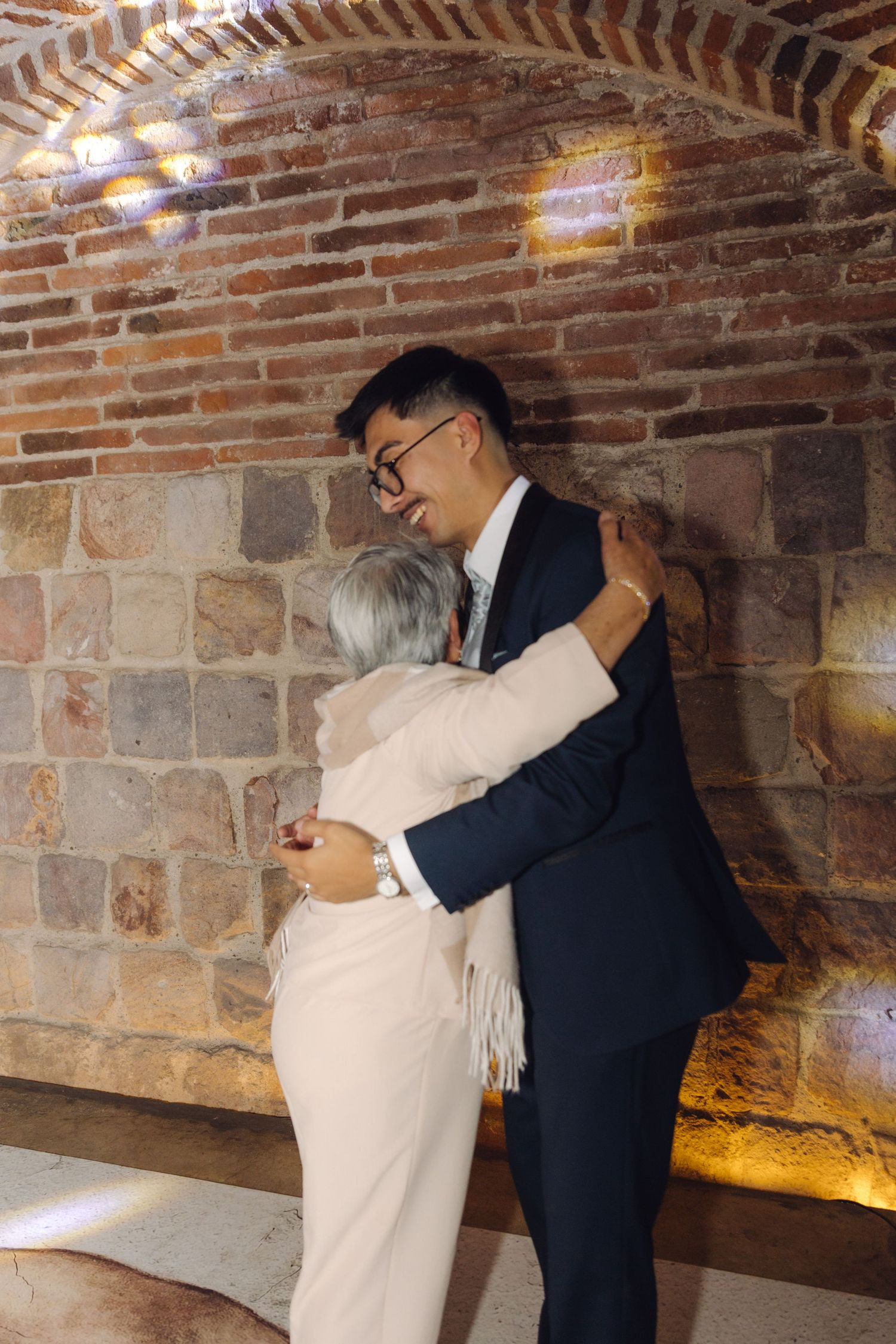 Groom in navy suit shares warm embrace with older woman against brick wall.