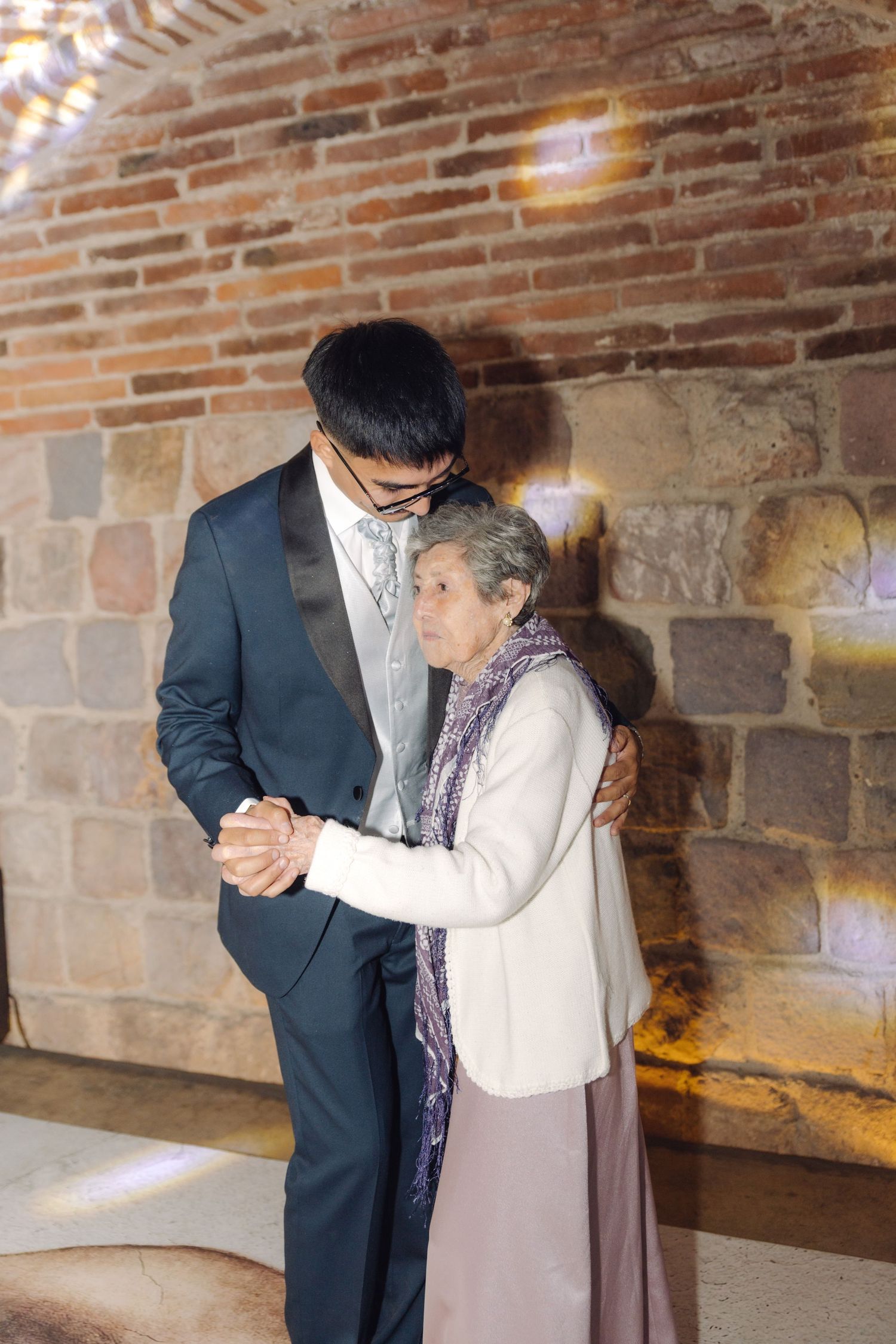 Groom in tuxedo shares tender dance moment with elderly guest at wedding reception.