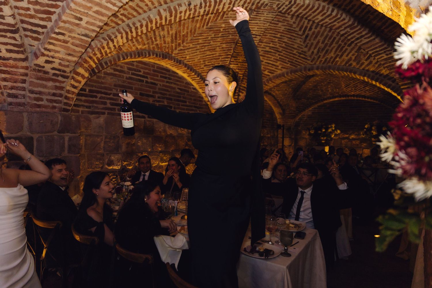 Woman in black dress celebrates joyfully holding wine bottle at elegant wedding reception in vaulted brick venue.