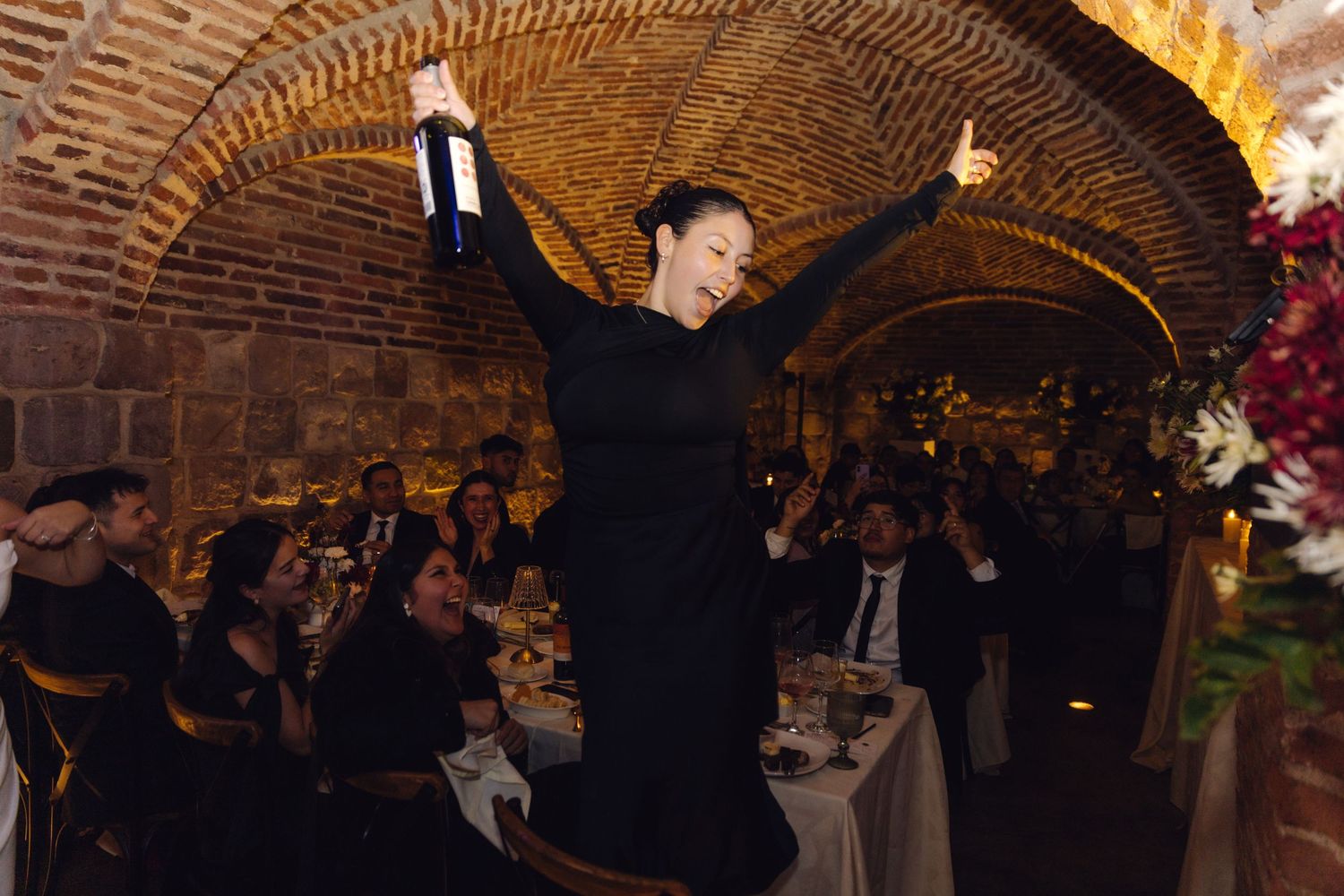 Elegant woman in black dress holds wine bottle aloft at candlelit wedding reception in historic brick arched venue.