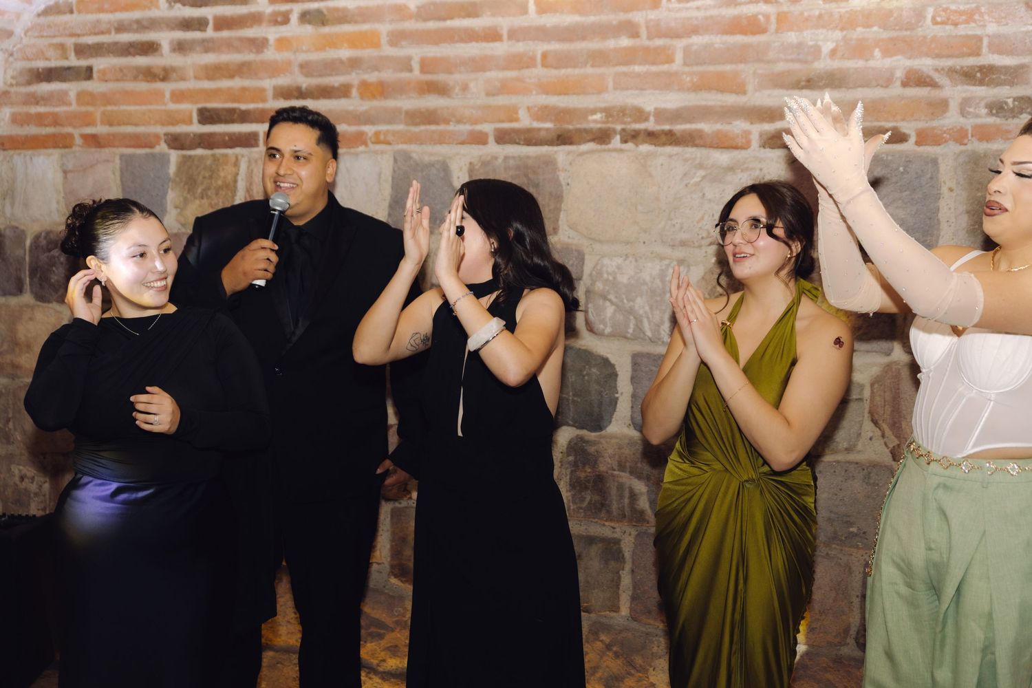 Group of wedding guests in formal attire laugh and clap while man speaks into microphone at reception.