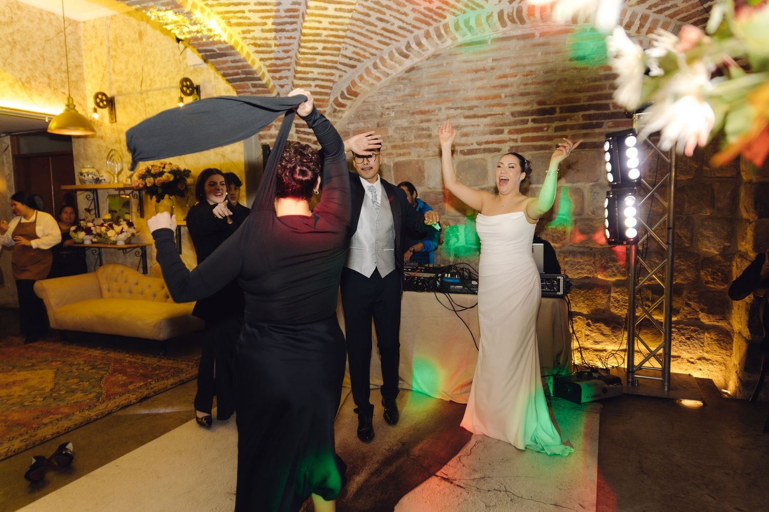 Bride in white gown dances with masked guest under colorful lights at lively wedding reception in brick venue.