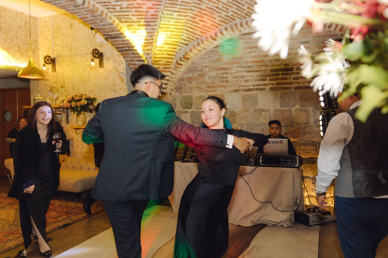 Couple dances energetically at wedding reception in rustic brick arched venue with guests watching nearby.