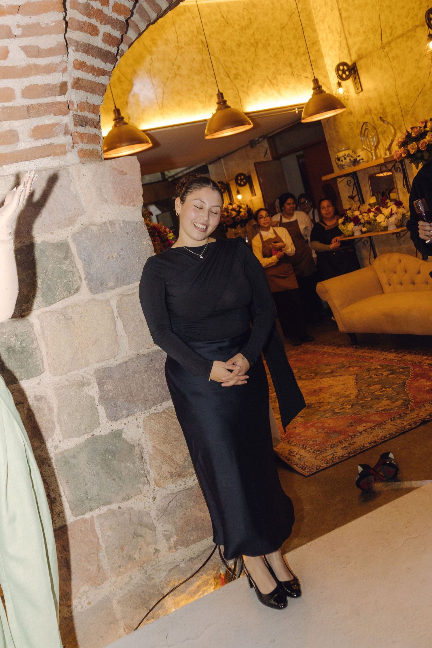 Elegant woman in black dress smiles while leaning against stone pillar in warmly lit wedding venue.