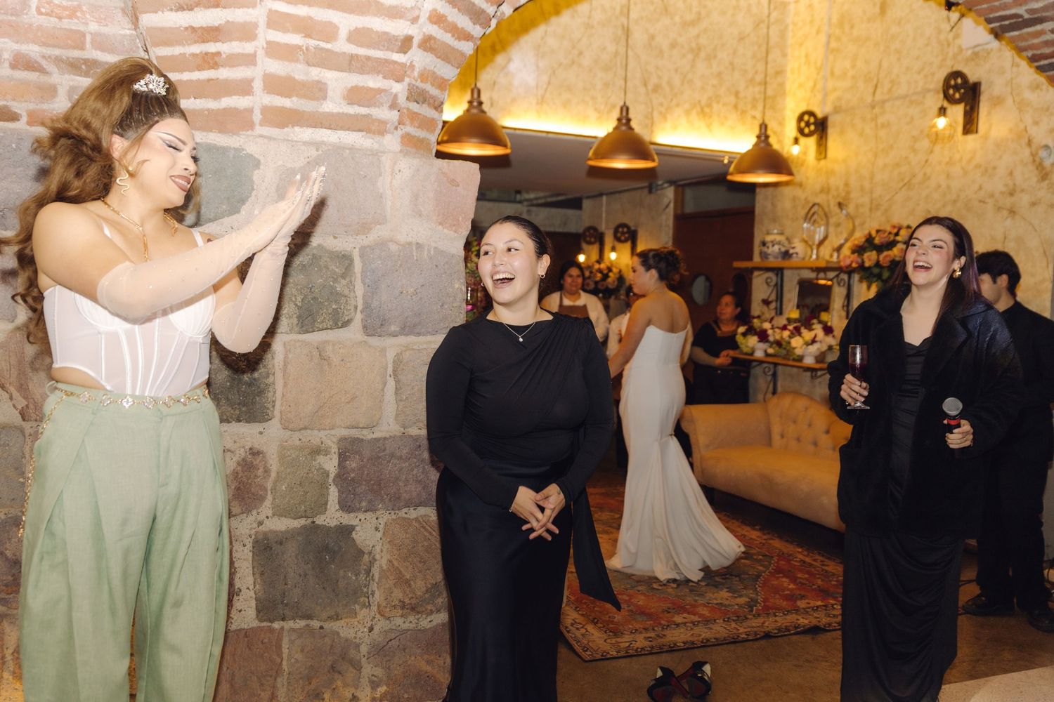 Three women in formal attire laugh and celebrate together at a wedding reception in a rustic brick venue.