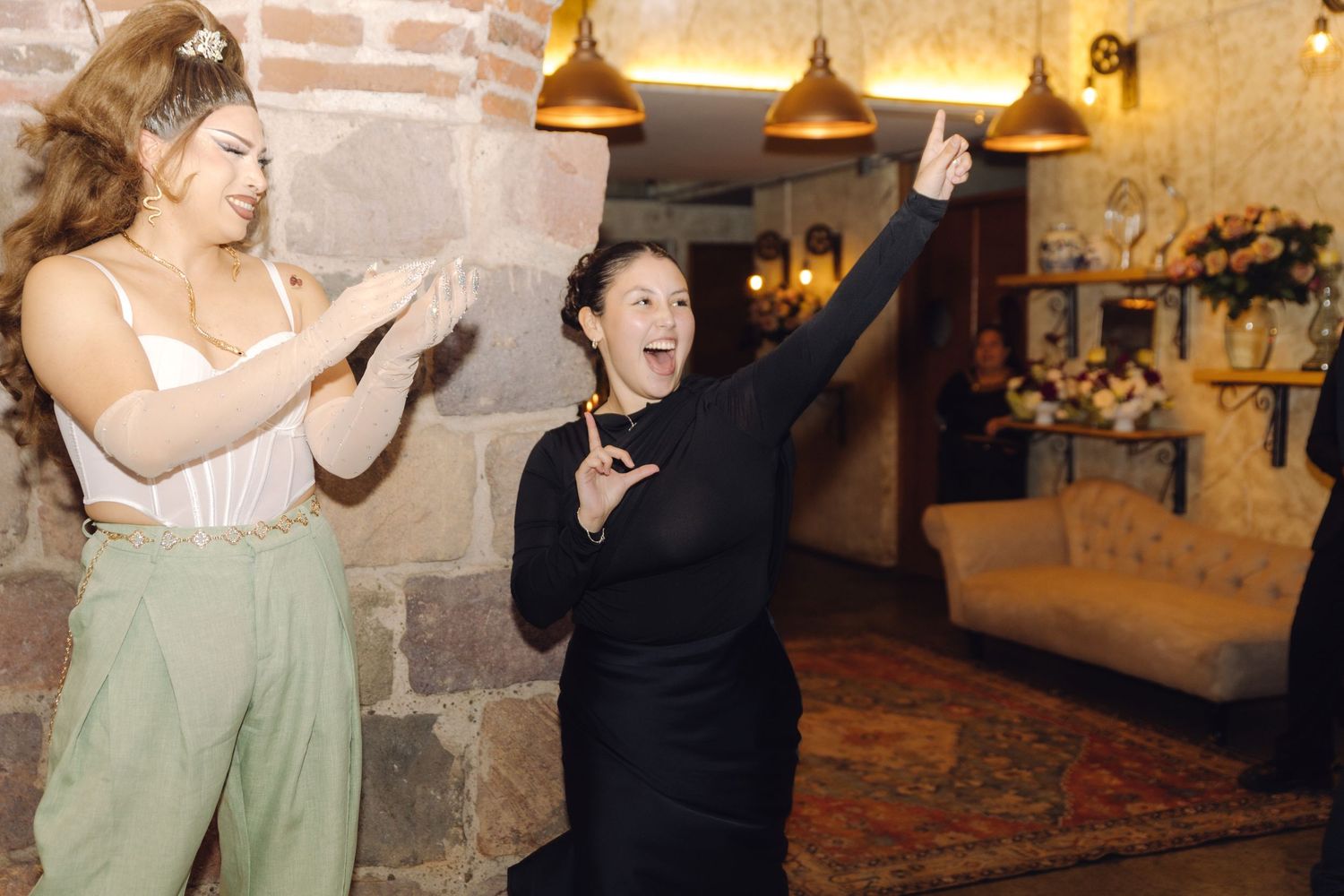 Two women laugh and dance joyfully at a wedding reception in a warmly lit rustic stone venue.