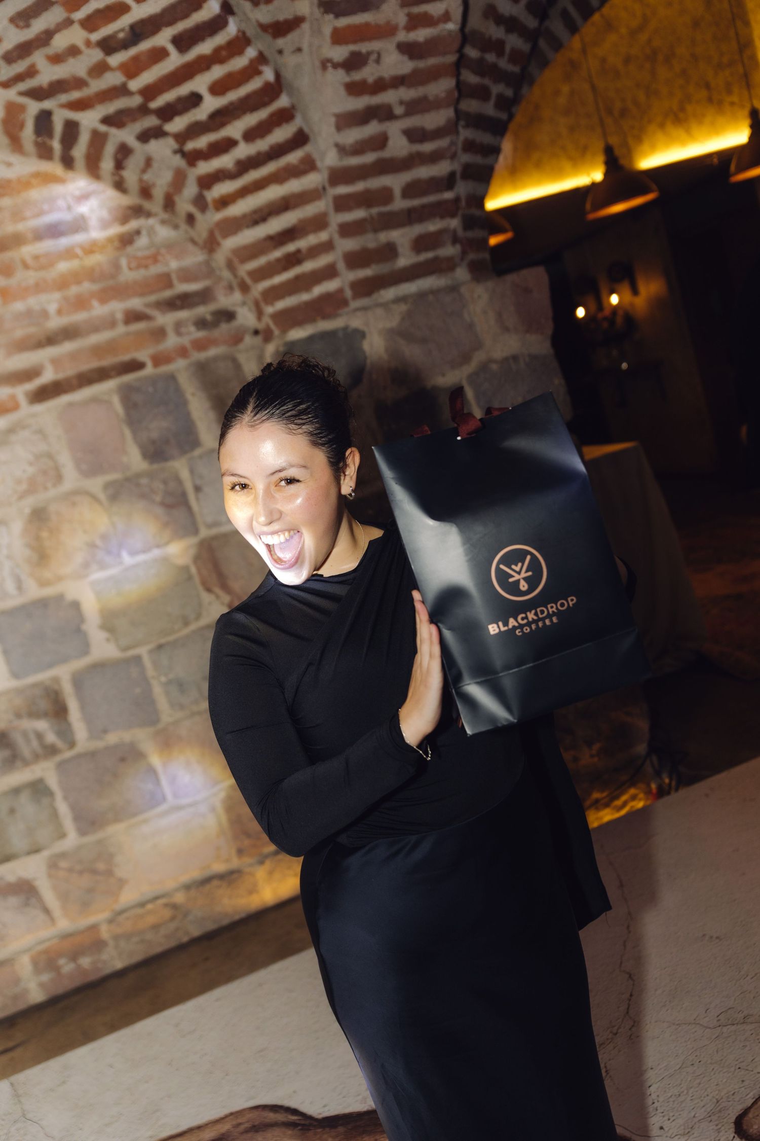 Woman in black dress poses playfully with a Black Drop coffee branded gift bag at a wedding reception.