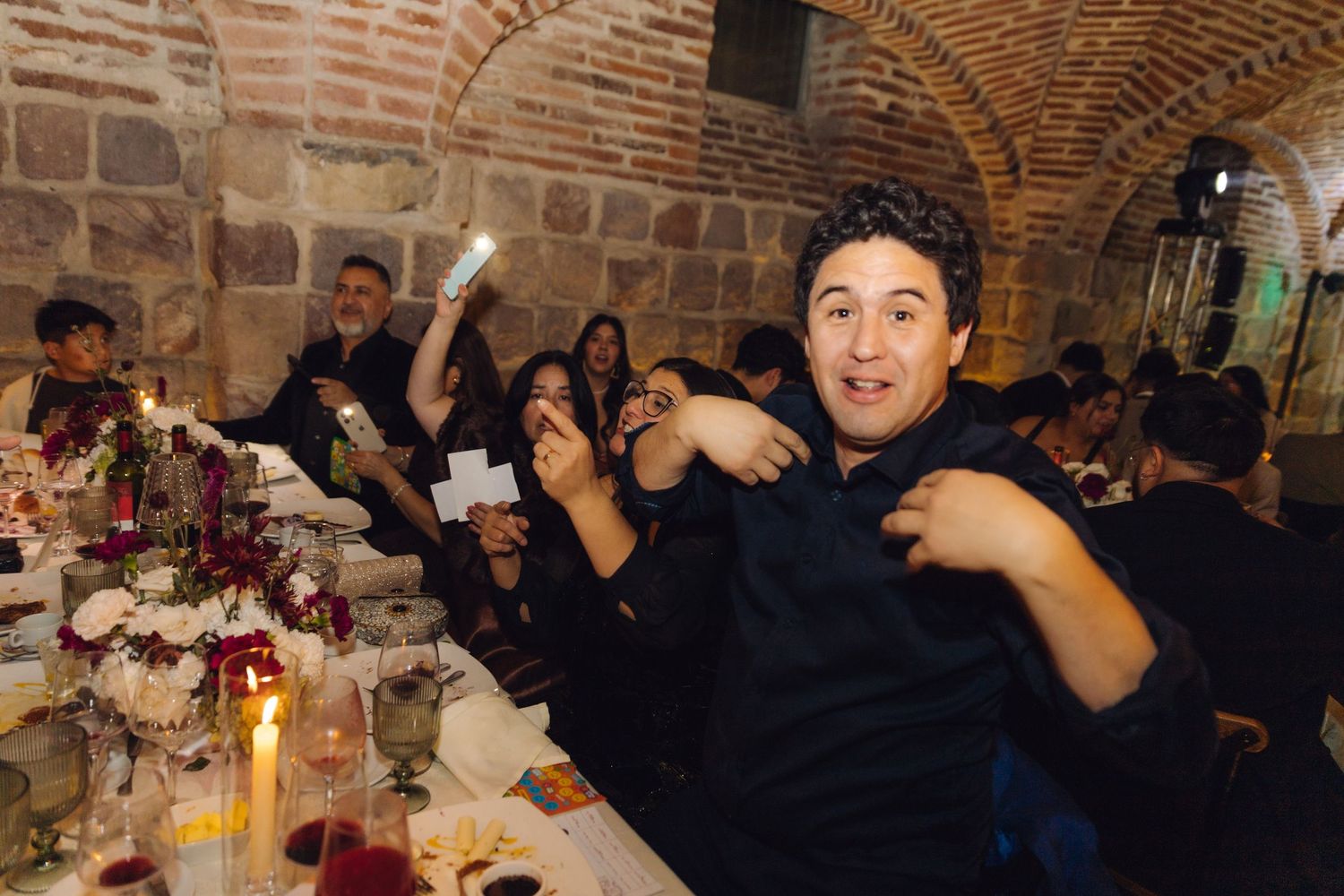 Guests celebrating at a wedding reception dinner in a rustic brick venue with candlelit tables and floral centerpieces.