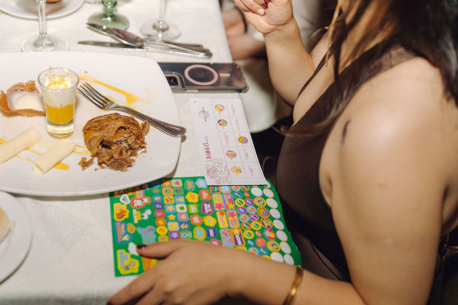 Wedding guest playing bingo at a reception dinner table with food, drinks, and colorful bingo cards.