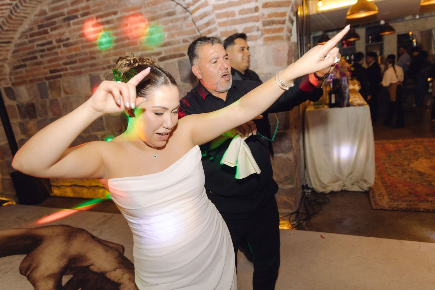 Bride in white gown dancing energetically with a guest at a wedding reception in a stone-walled venue.