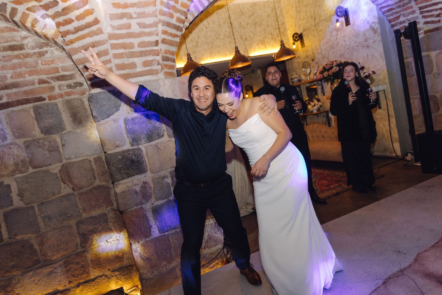 Bride and groom celebrating joyfully at their wedding reception in a warmly lit rustic brick venue.