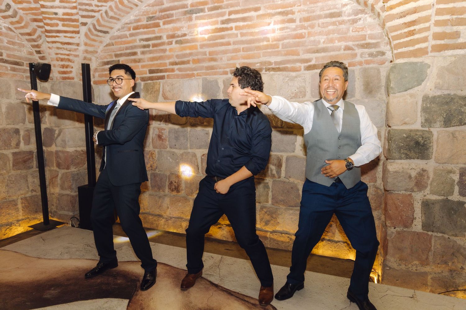 Three men striking a fun dancing pose together at a wedding reception in a brick-walled venue.