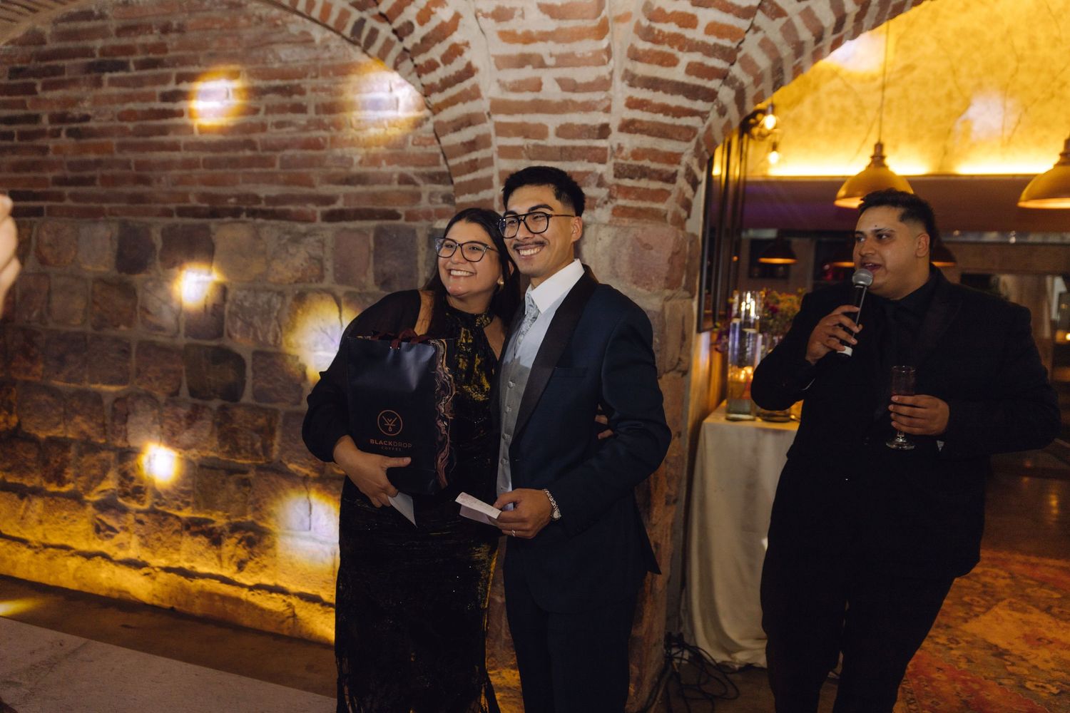 Three wedding guests posing together in a warmly lit rustic brick venue during a reception celebration.