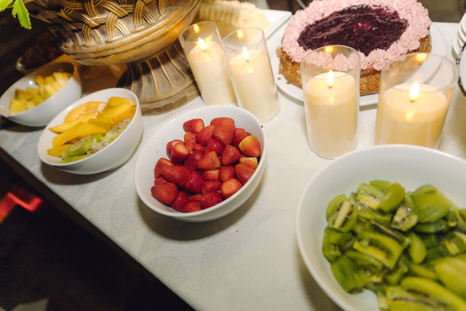 Wedding dessert table featuring fresh strawberries, kiwi, mango, and a decorated cake with lit candles.