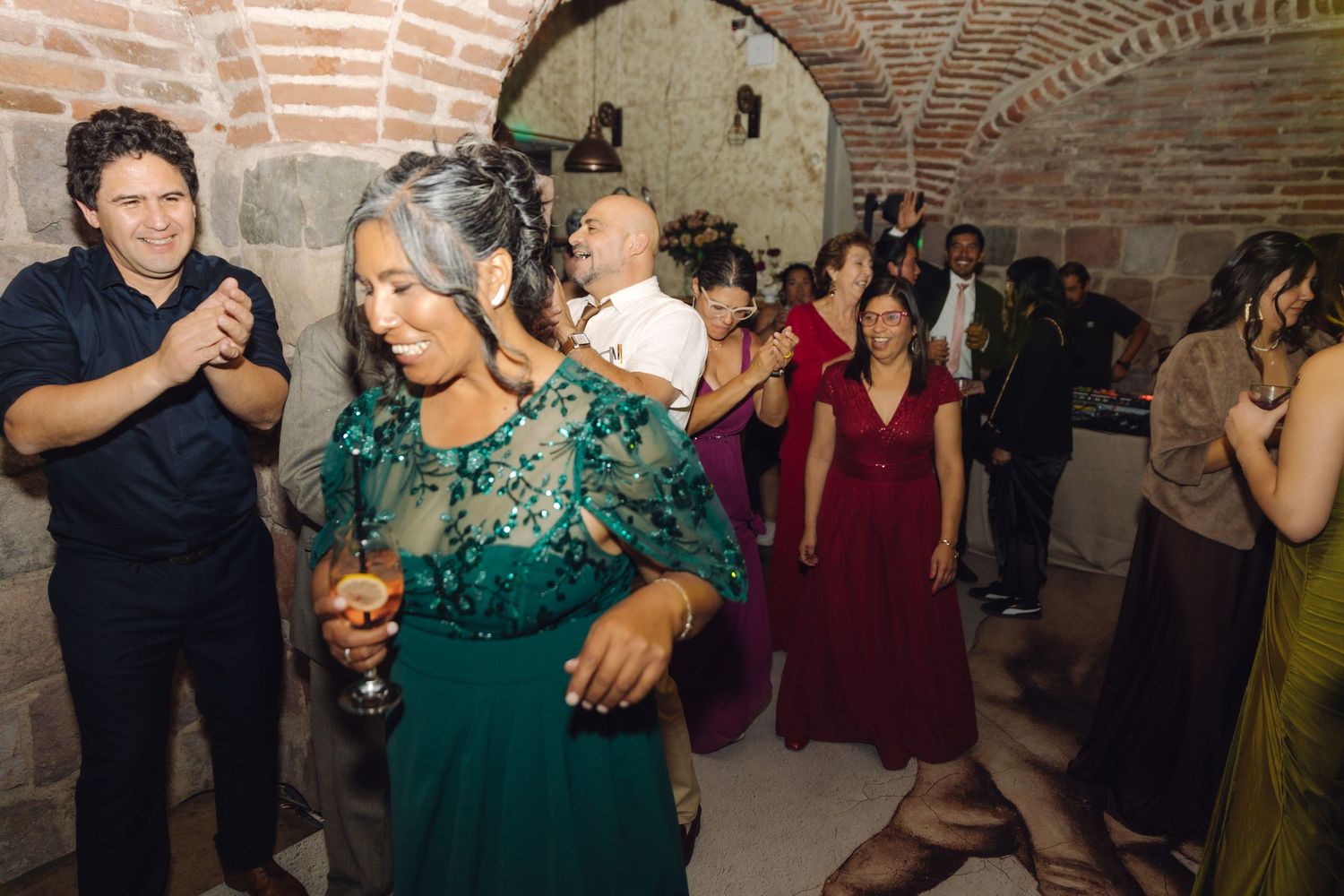Elegantly dressed guests dancing at a festive wedding reception in a beautiful stone arched venue.