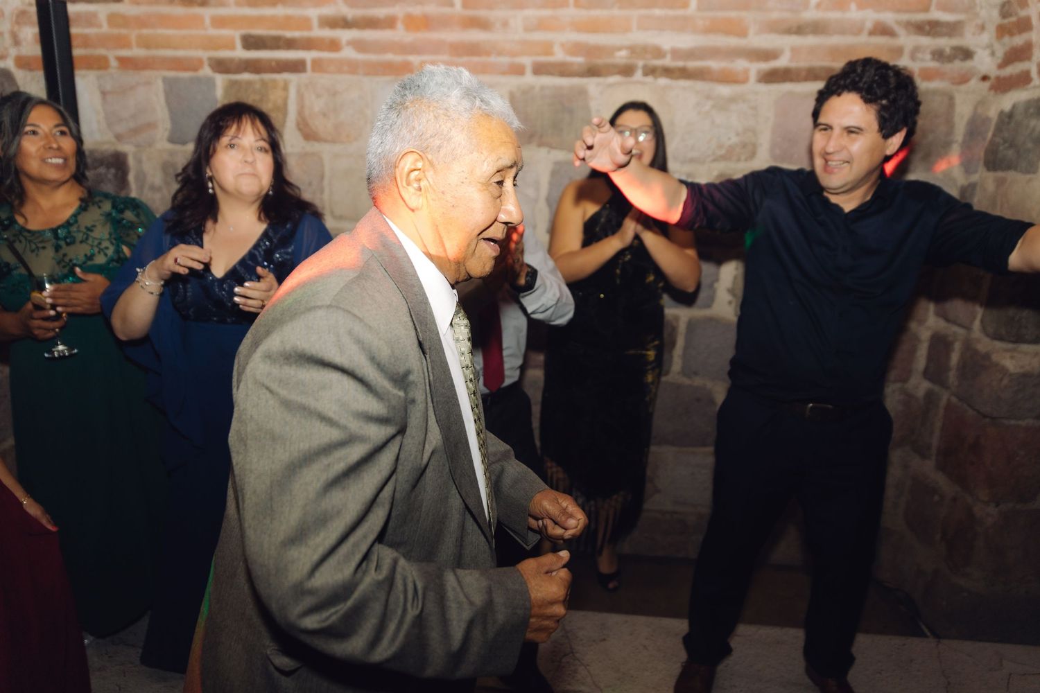 Elderly man dancing energetically at a wedding reception while guests cheer in a stone-walled venue.