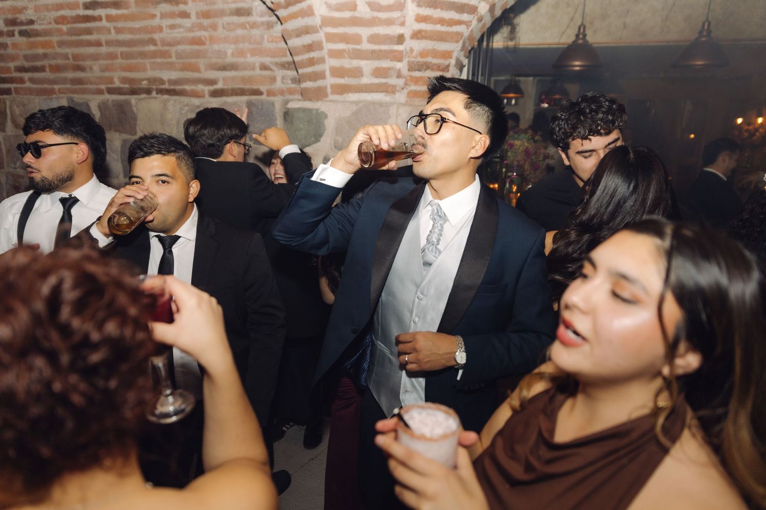 Young man in tuxedo drinking at a lively wedding reception party in a rustic brick venue.