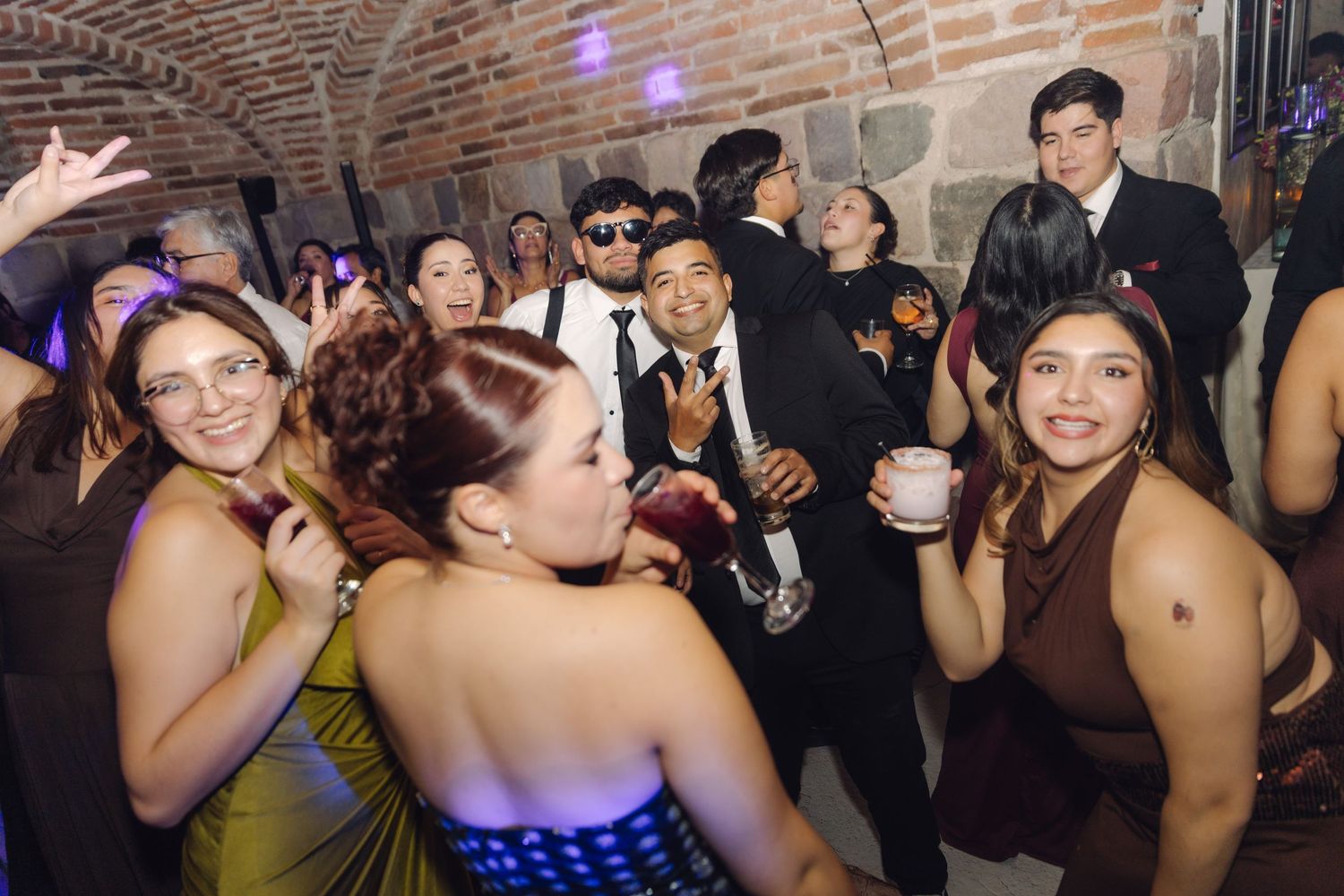 Young guests laughing and dancing together at a vibrant wedding reception in a stone-walled venue.