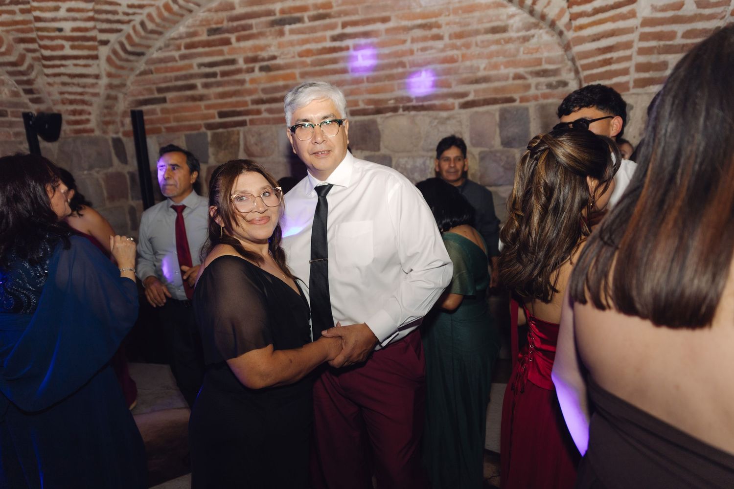 Couple dancing together at an elegant wedding reception in a rustic brick venue with purple lighting.