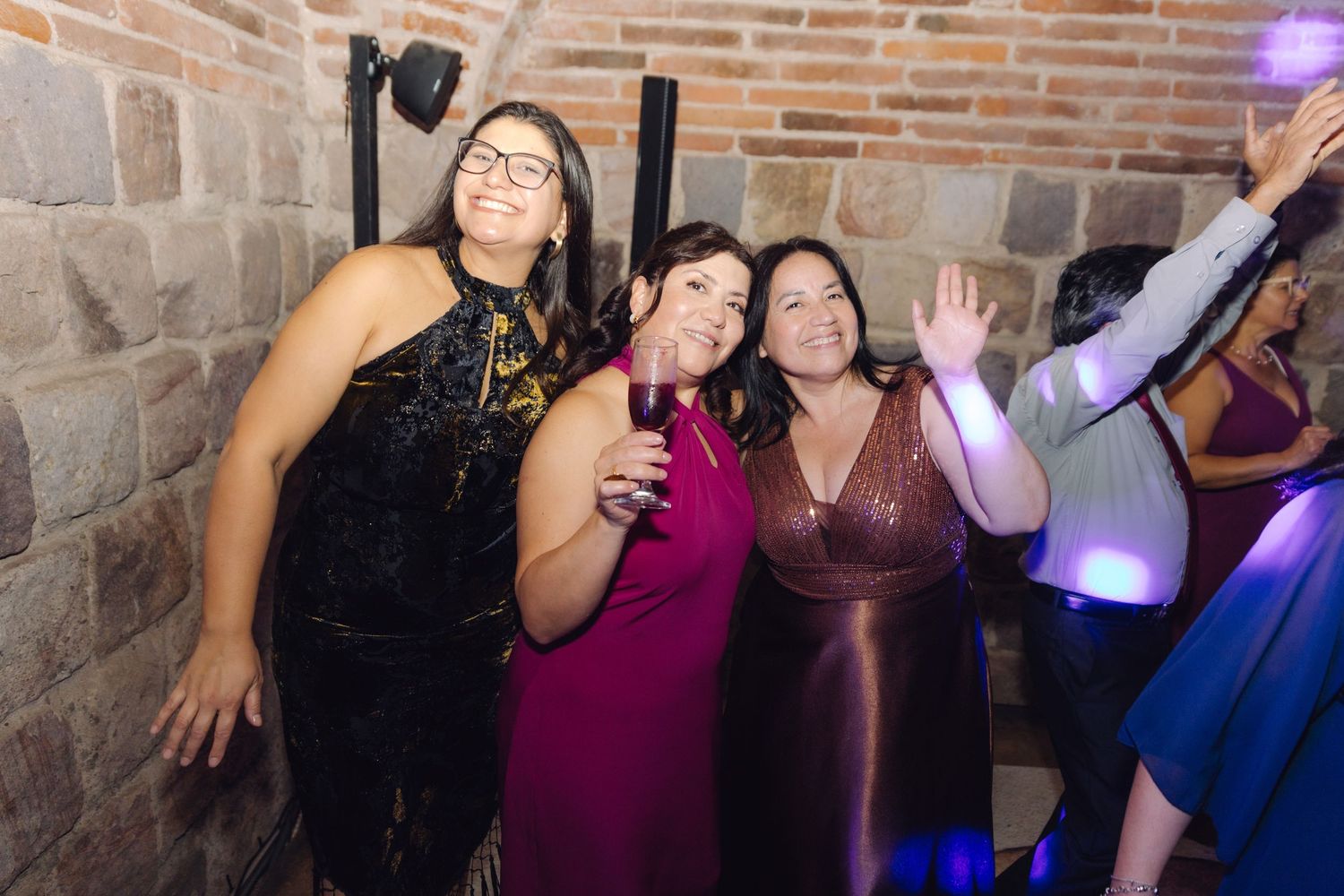 Three women in formal dresses celebrating and dancing at a wedding reception against a brick wall.