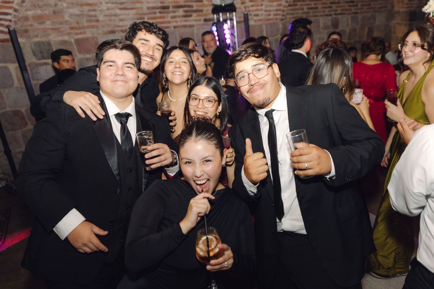 Group of young guests in black suits and formal wear posing with champagne at a wedding party.