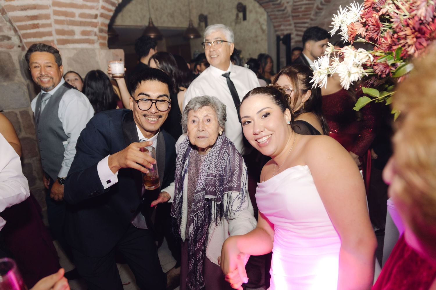 Bride in white dress posing with an elderly woman and guests at a warmly lit wedding reception.