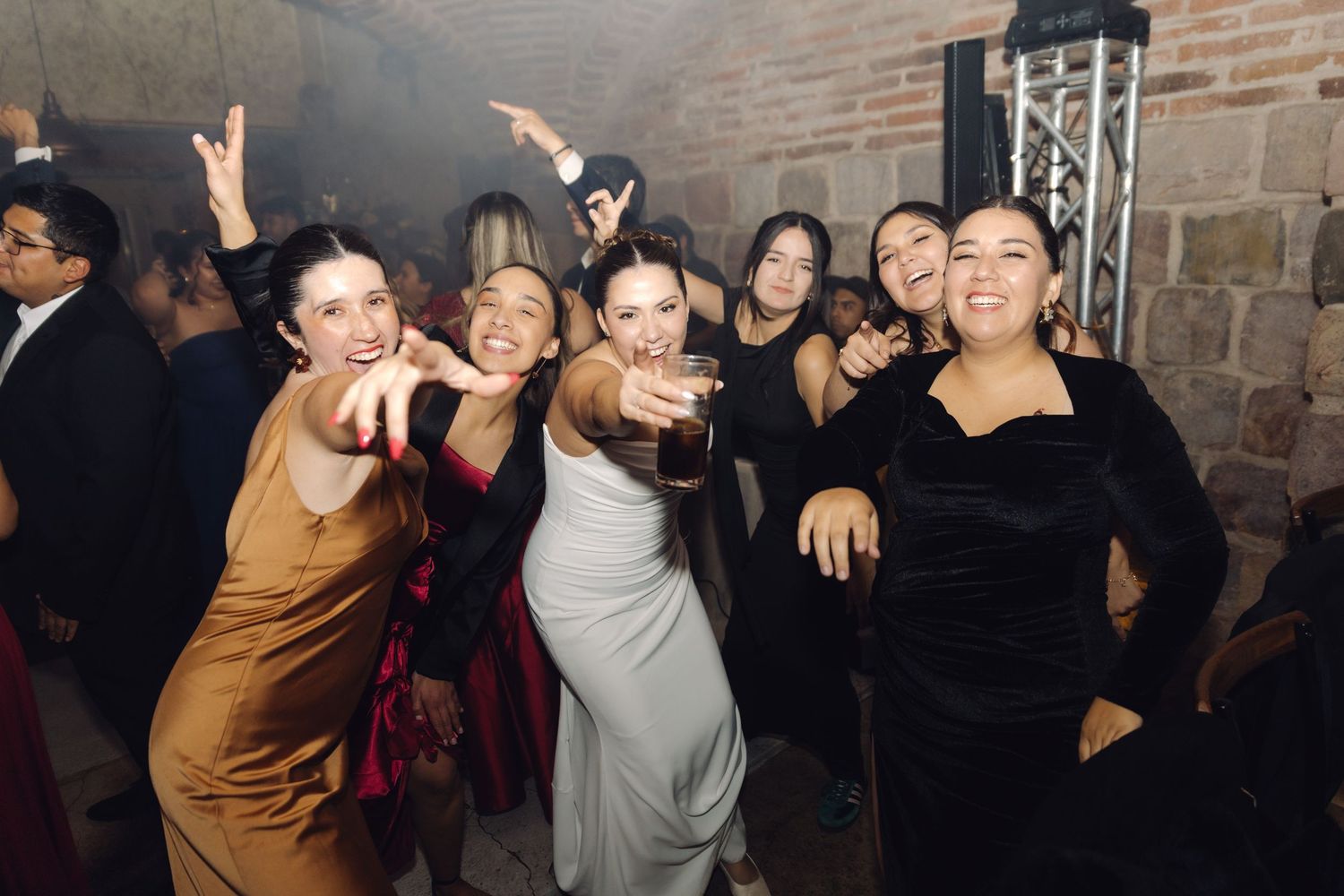 Group of women in elegant dresses dancing and pointing at the camera at a wedding celebration.