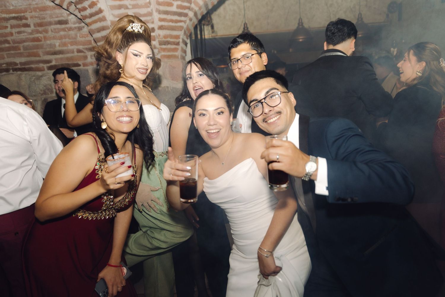 Bride in white dress surrounded by smiling guests holding drinks at a vibrant wedding reception.
