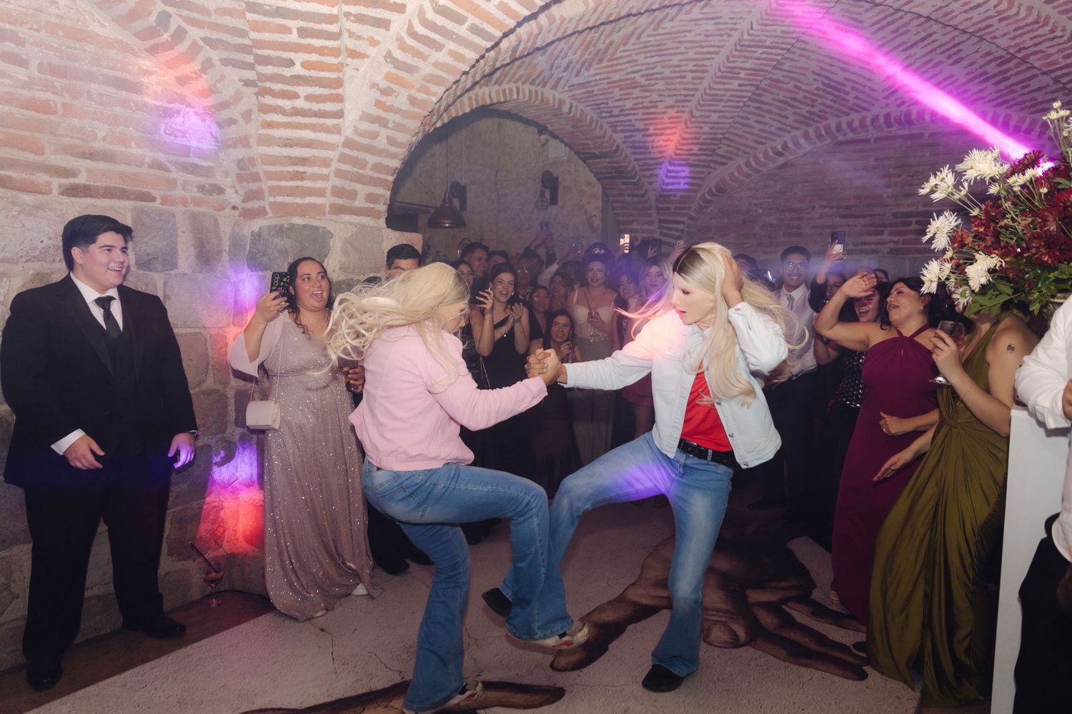 Two women in cowboy hats dance energetically at a wedding reception in a rustic brick venue.