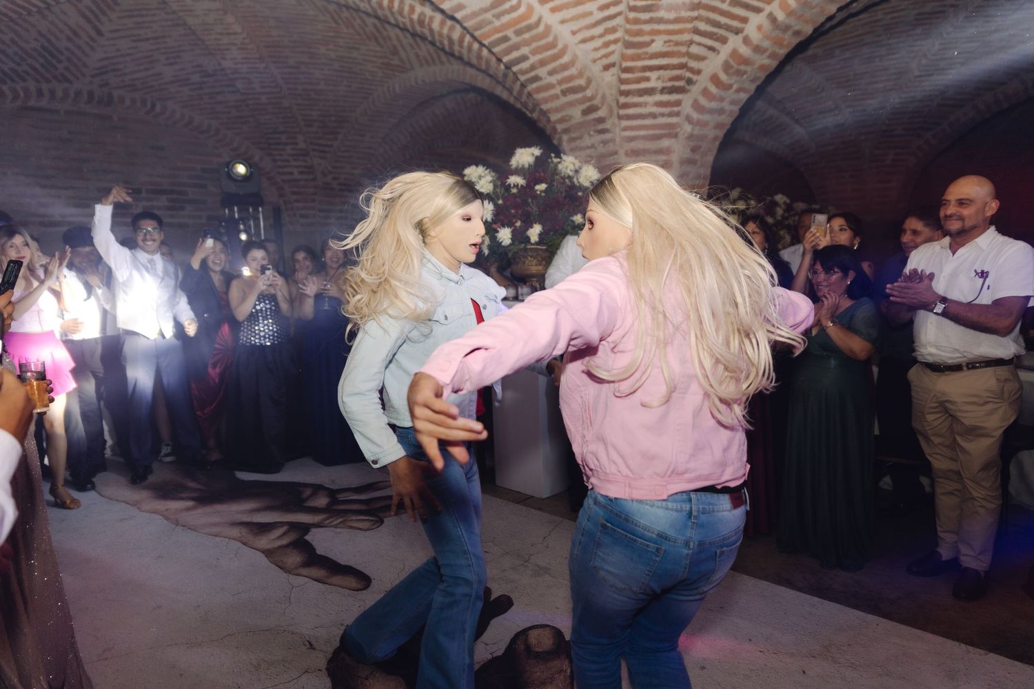 Two blonde women dance together energetically while guests applaud at a wedding reception.