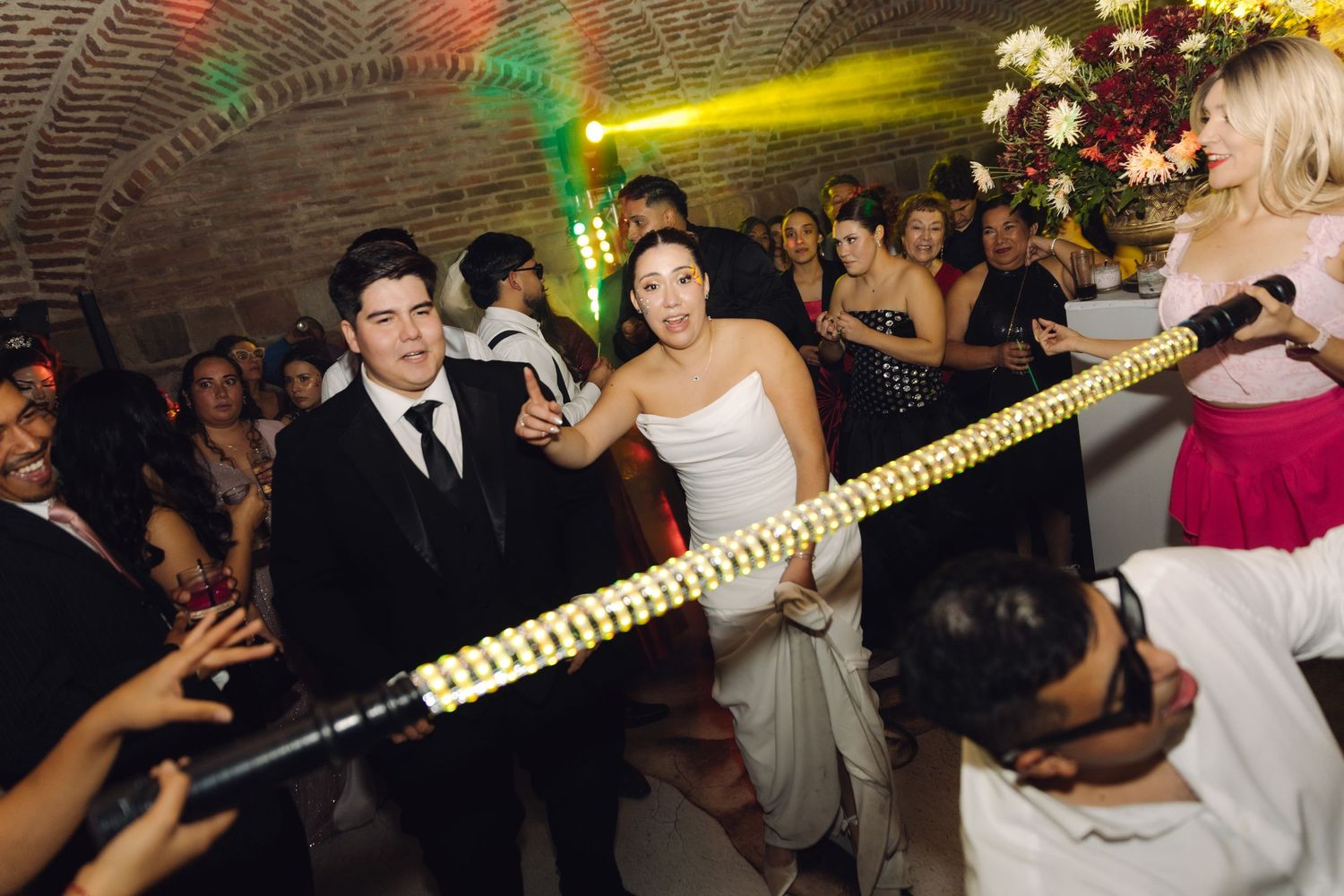 Newlyweds laugh while doing the limbo together under a lit bar at their wedding reception.