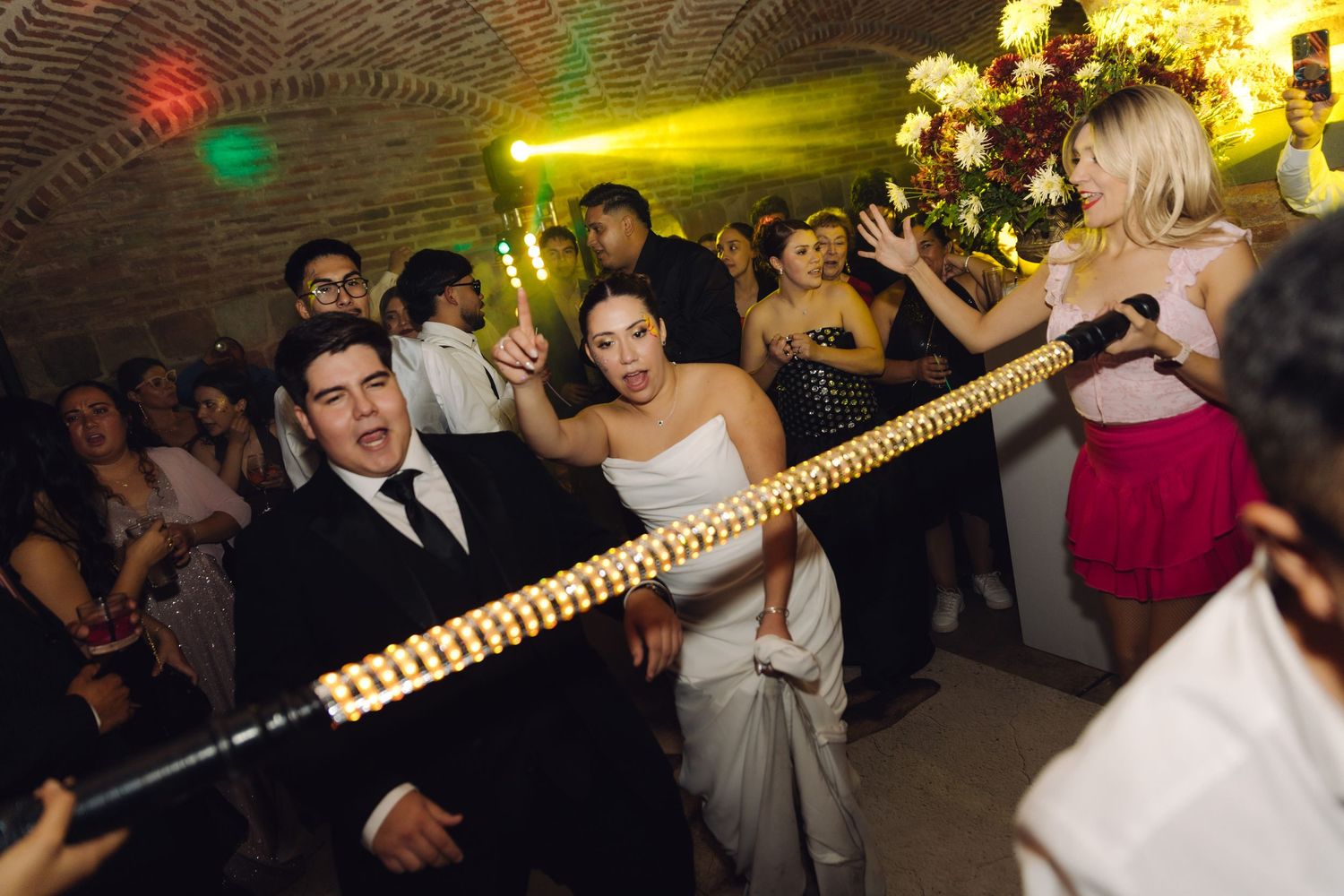 Bride in white gown and groom do the limbo under a glowing bar at their wedding.