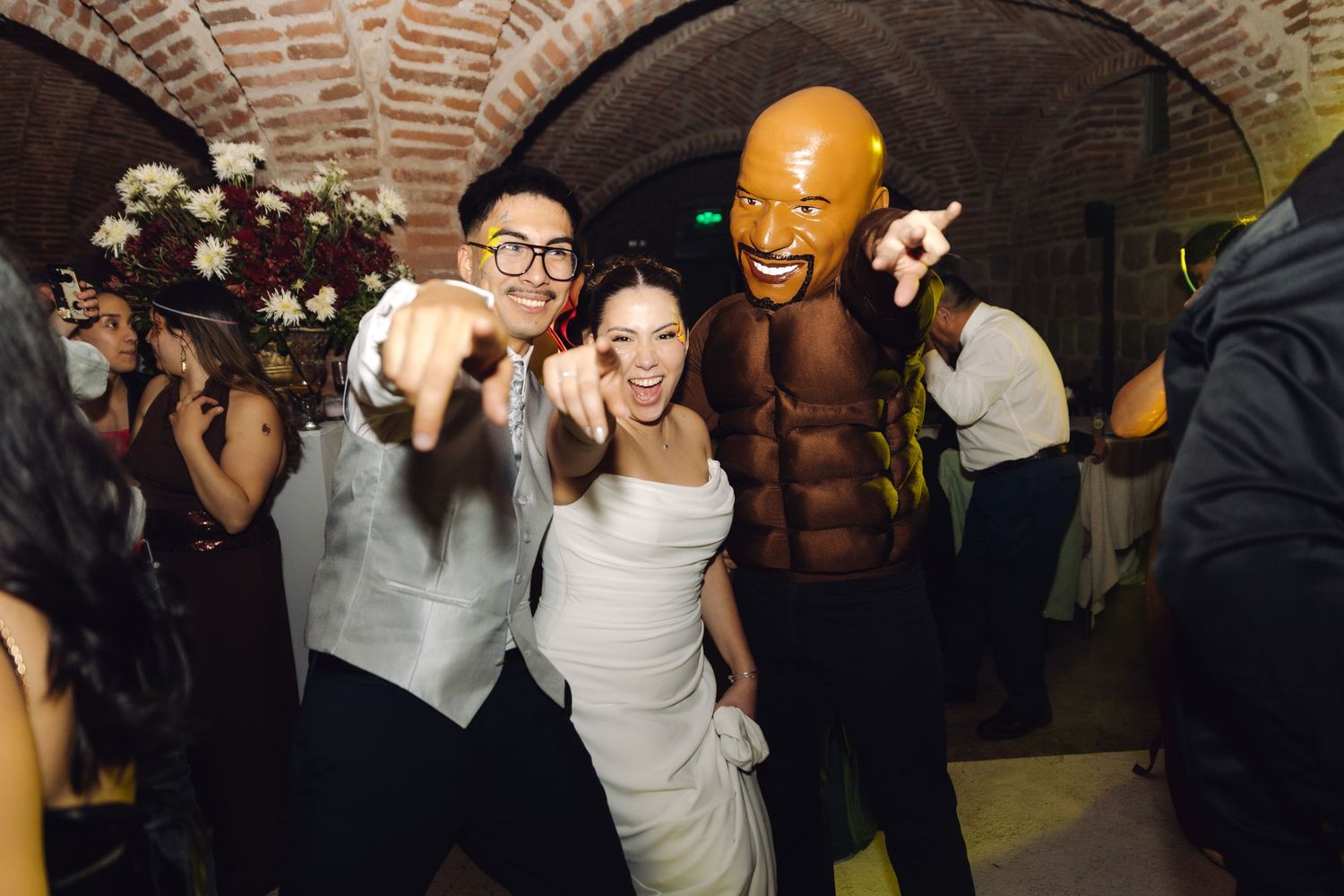 Bride and groom pose with costumed guest at lively wedding reception in brick arched venue.