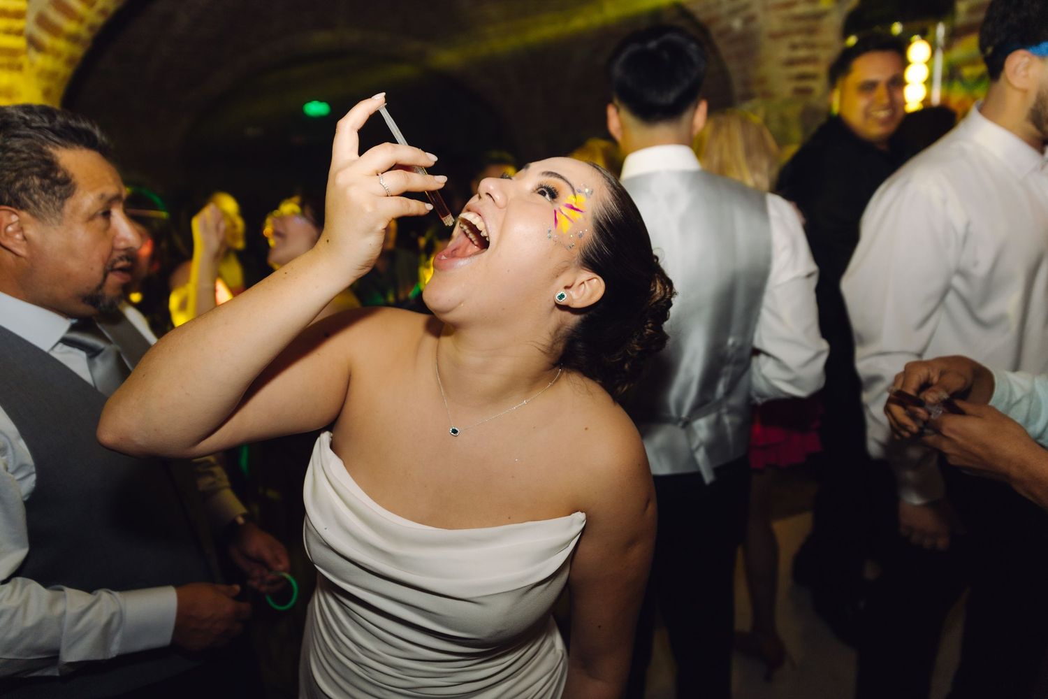 Joyful bride laughs while taking selfie on crowded wedding reception dance floor.