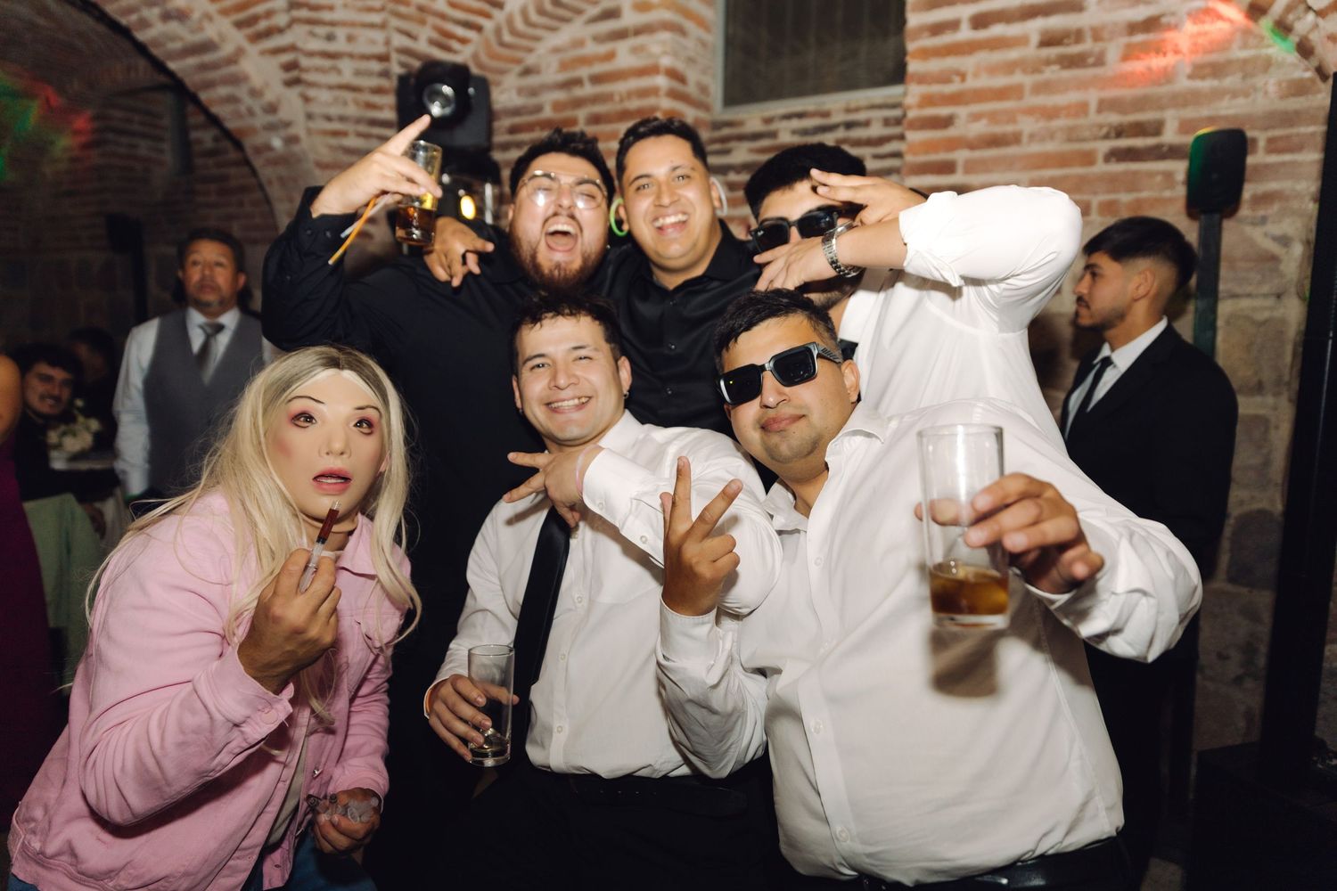 Group of wedding guests pose playfully with sunglasses and drinks at reception party.