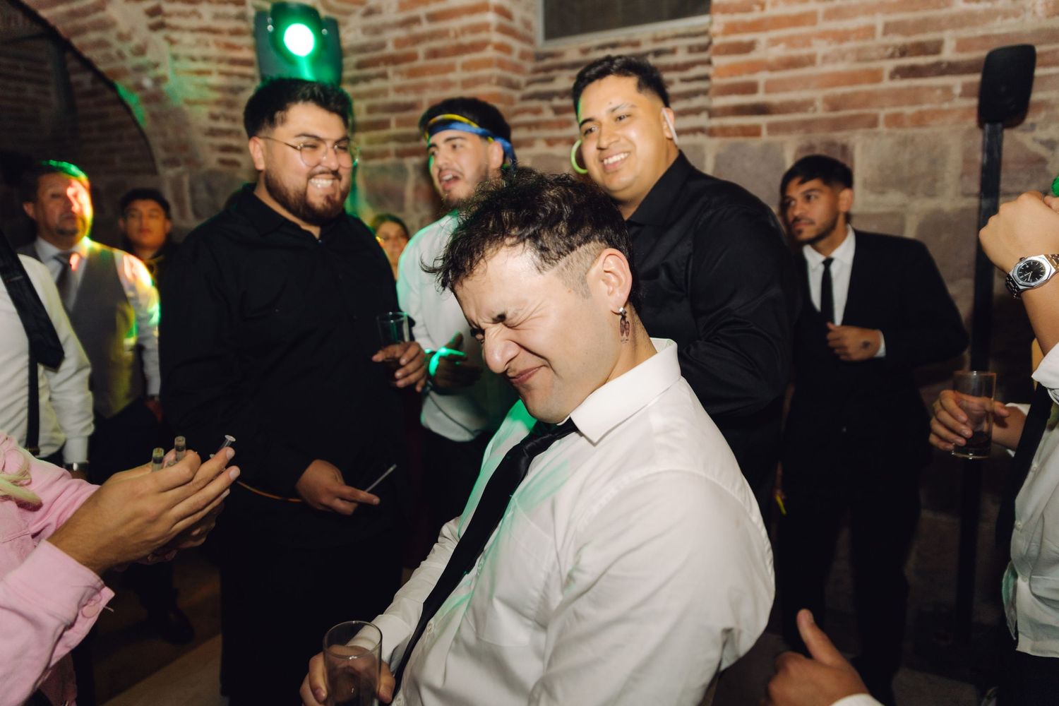Wedding guests cheer as man dances at lively reception inside brick arched venue.