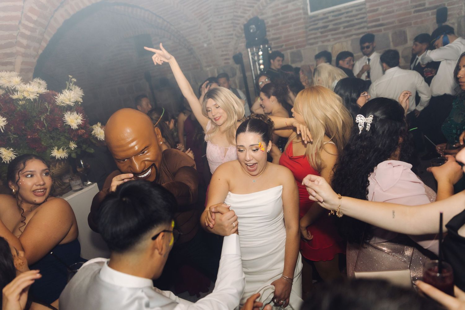 Bride dances joyfully with guests at crowded wedding reception in brick arched hall.
