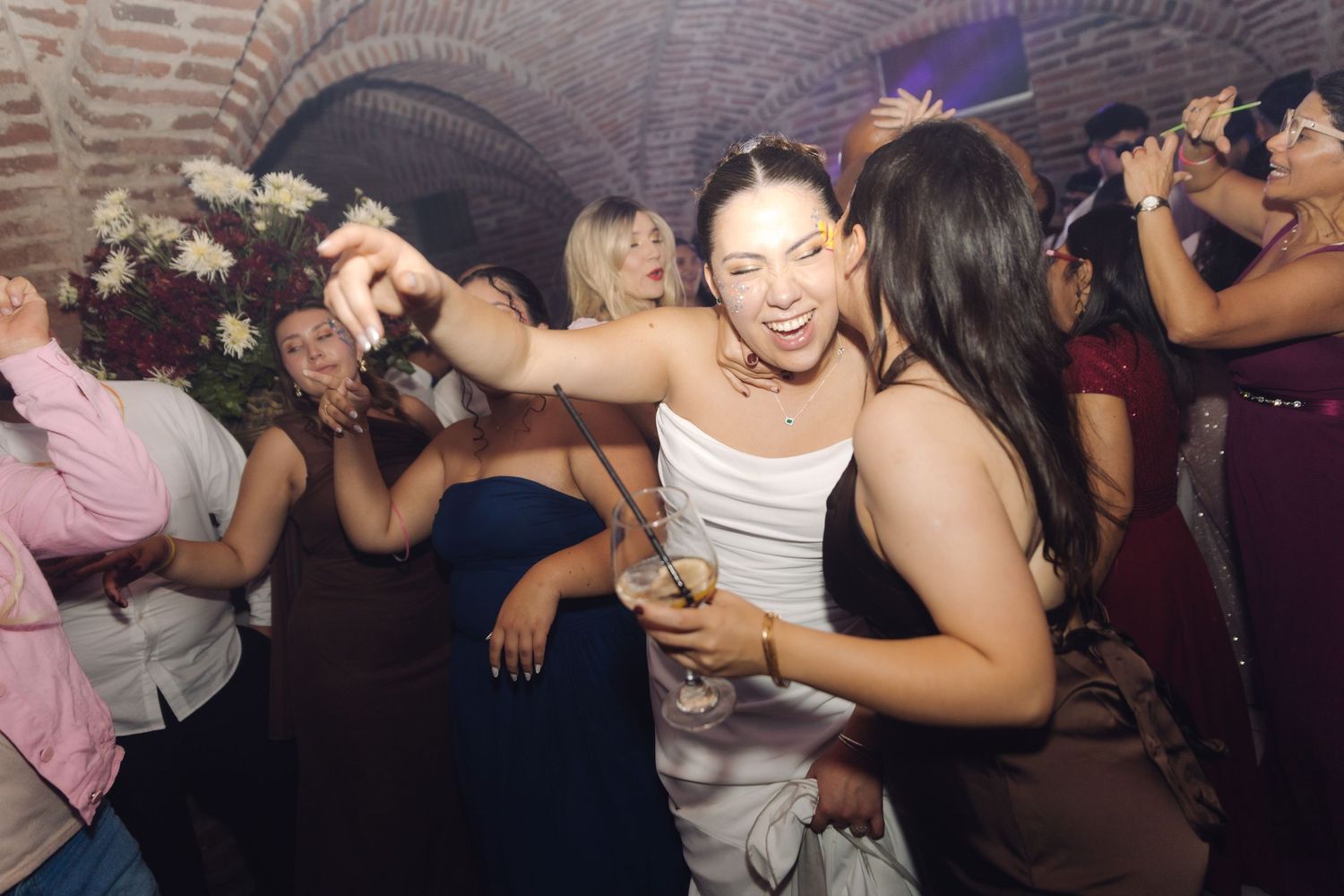 Smiling bride dances with female guest while holding drink at wedding reception party.