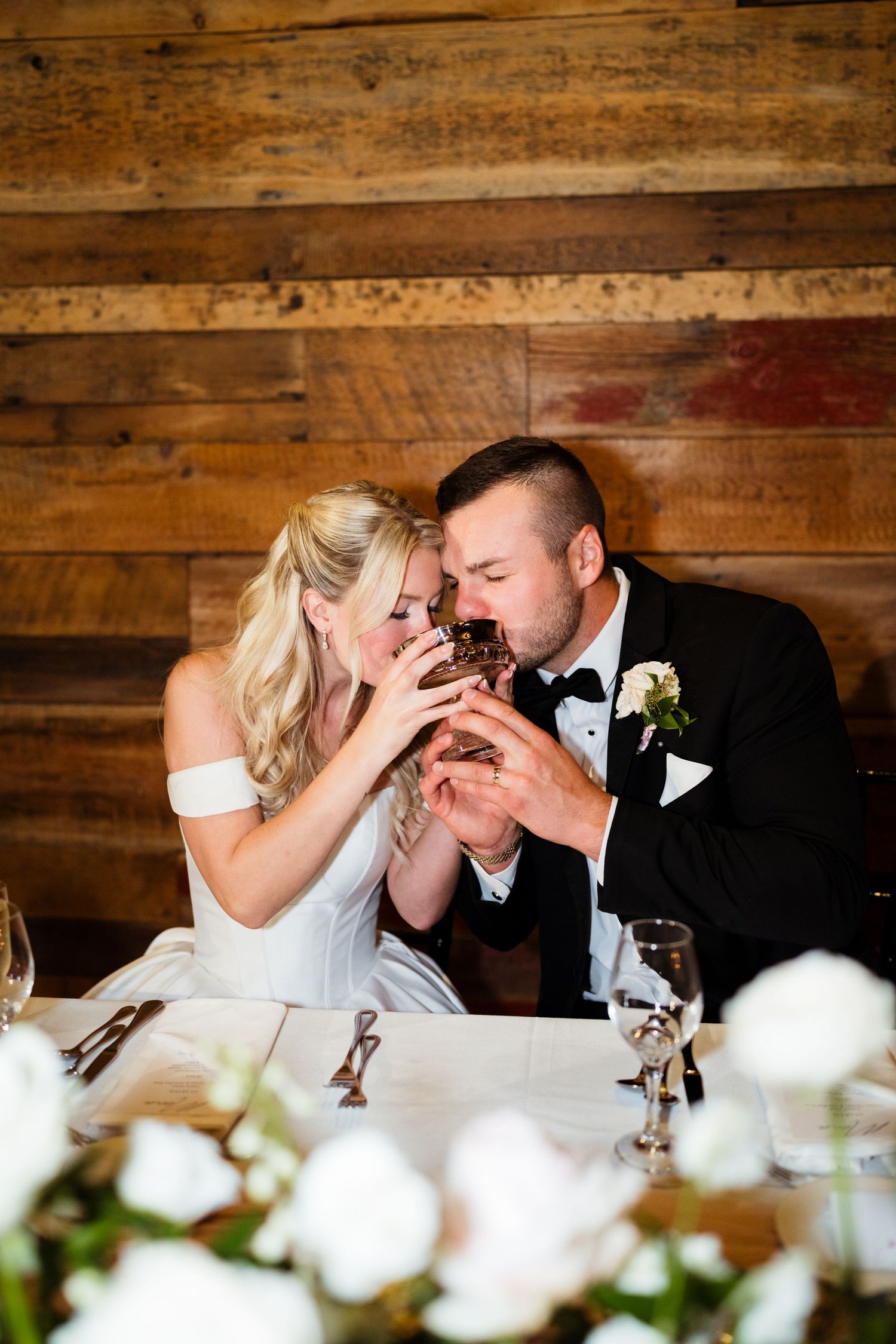 Bride and groom sharing wedding cake at rustic barn reception table with white floral centerpieces.